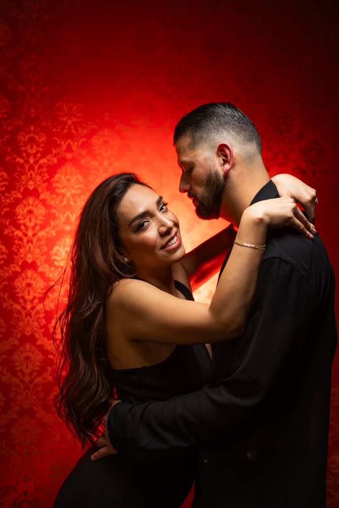Elegant couple sharing a playful moment against red patterned backdrop.