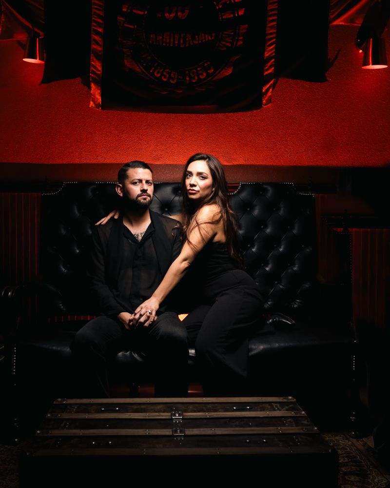 A couple poses intimately on a black leather couch in dramatic lighting.