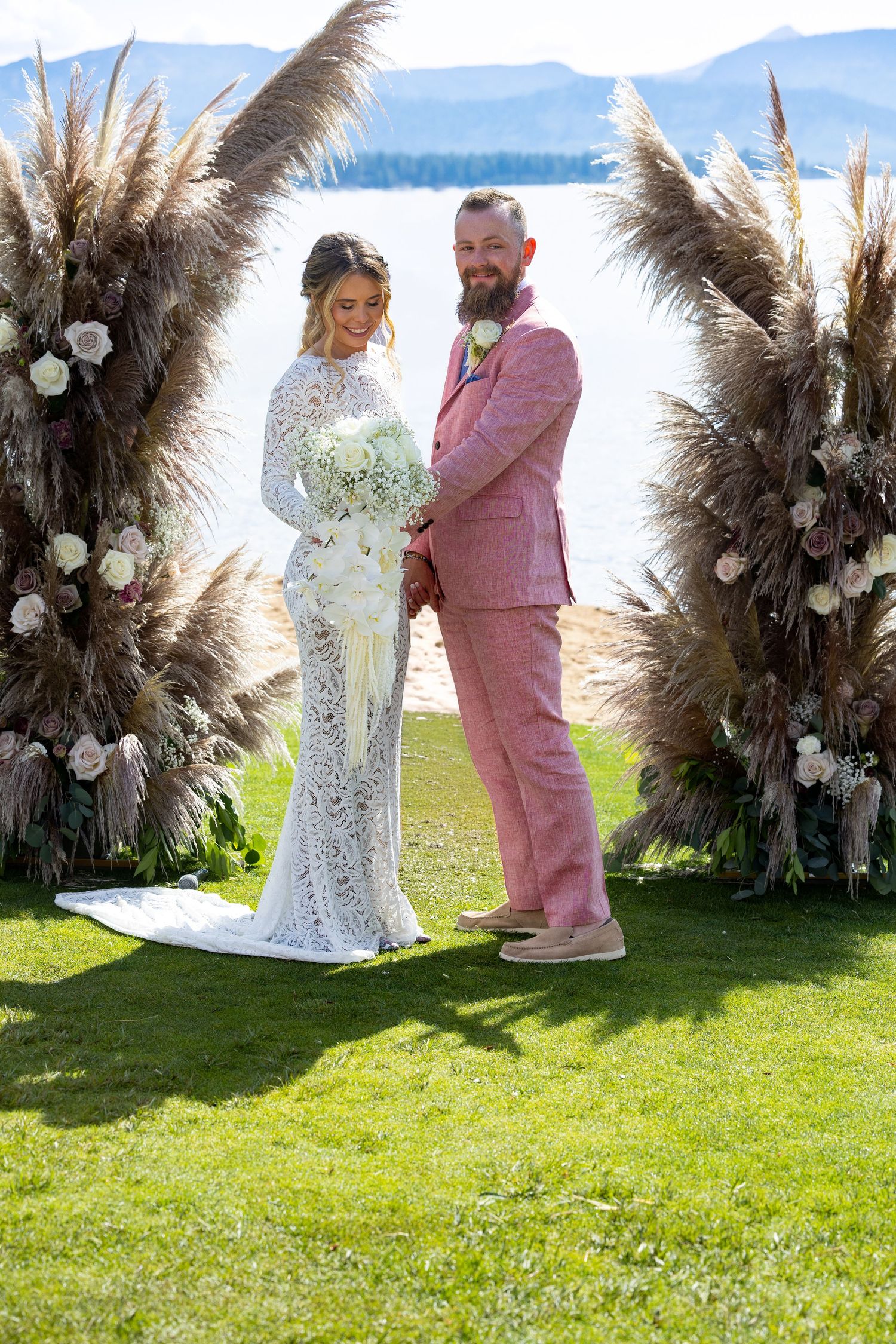A wedding couple poses under a dramatic pampas grass arch by a lakeside in elegant pink suit and white lace gown.