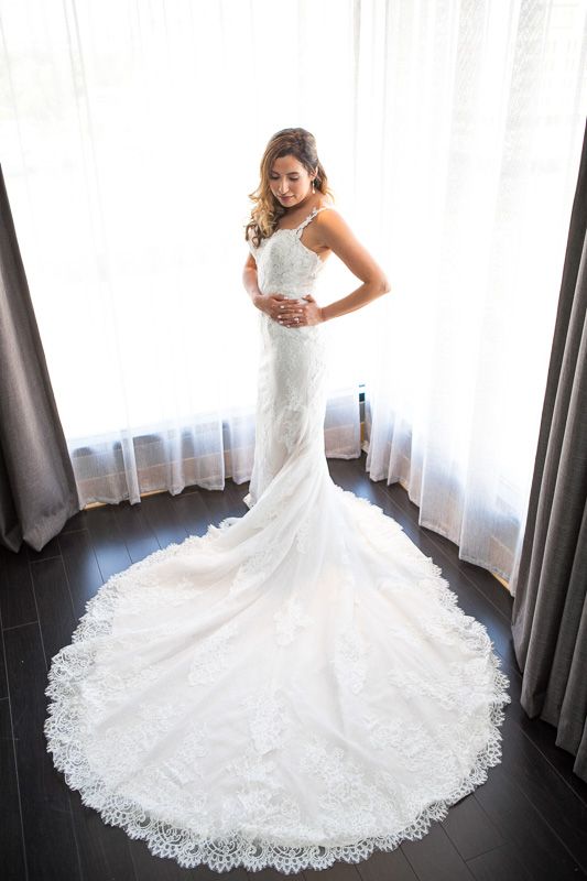 A stunning lace wedding gown with dramatic cathedral train displayed in front of sheer white curtains.
