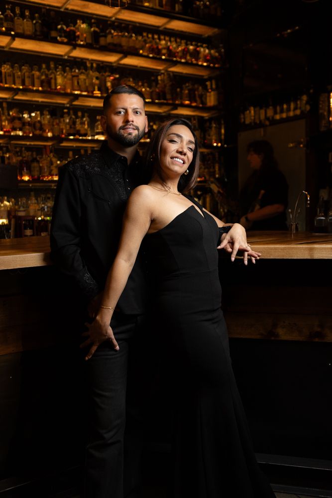 Elegant couple in black attire posing together against bar backdrop with ambient lighting.