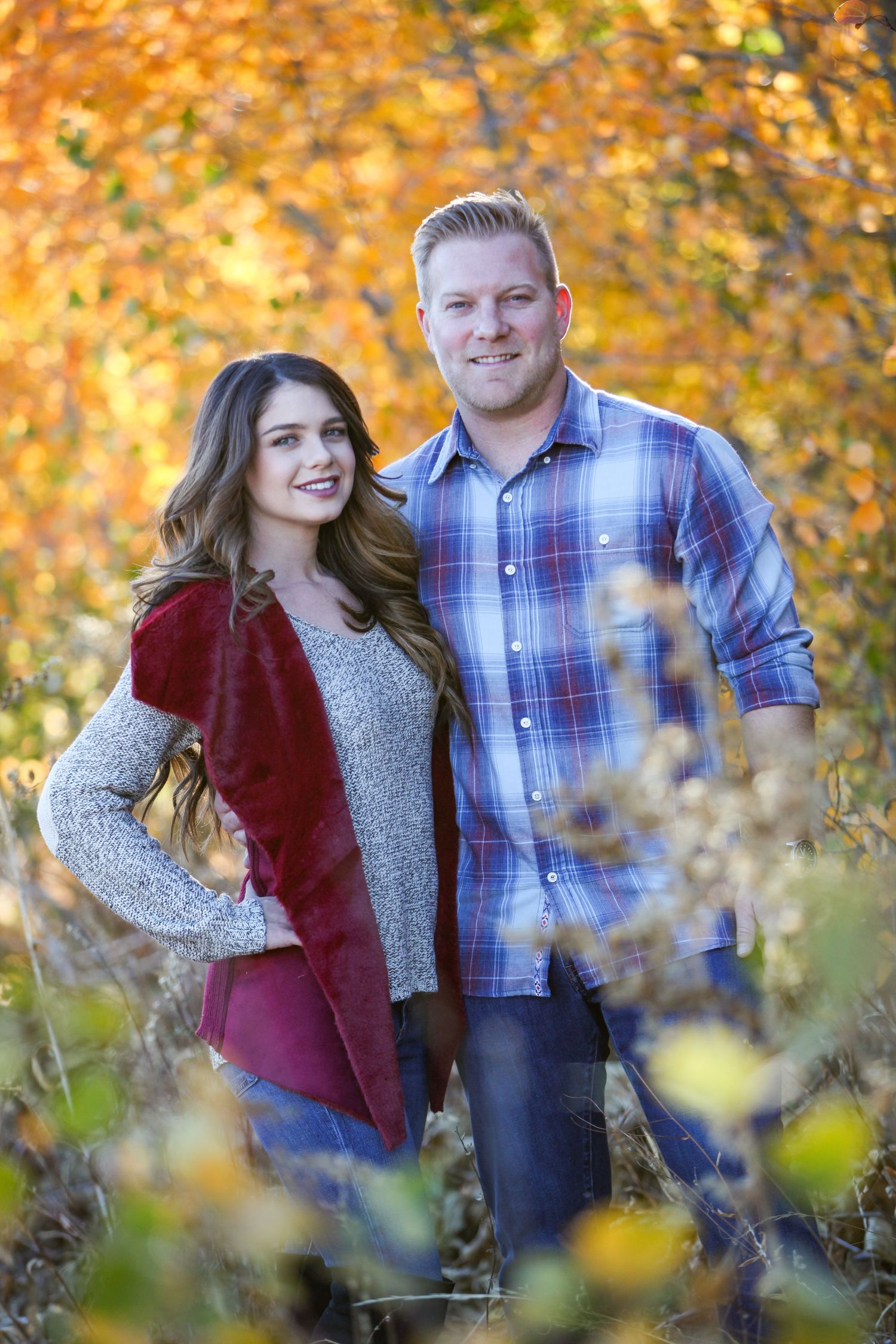 Couple in casual fall attire poses among golden autumn leaves.