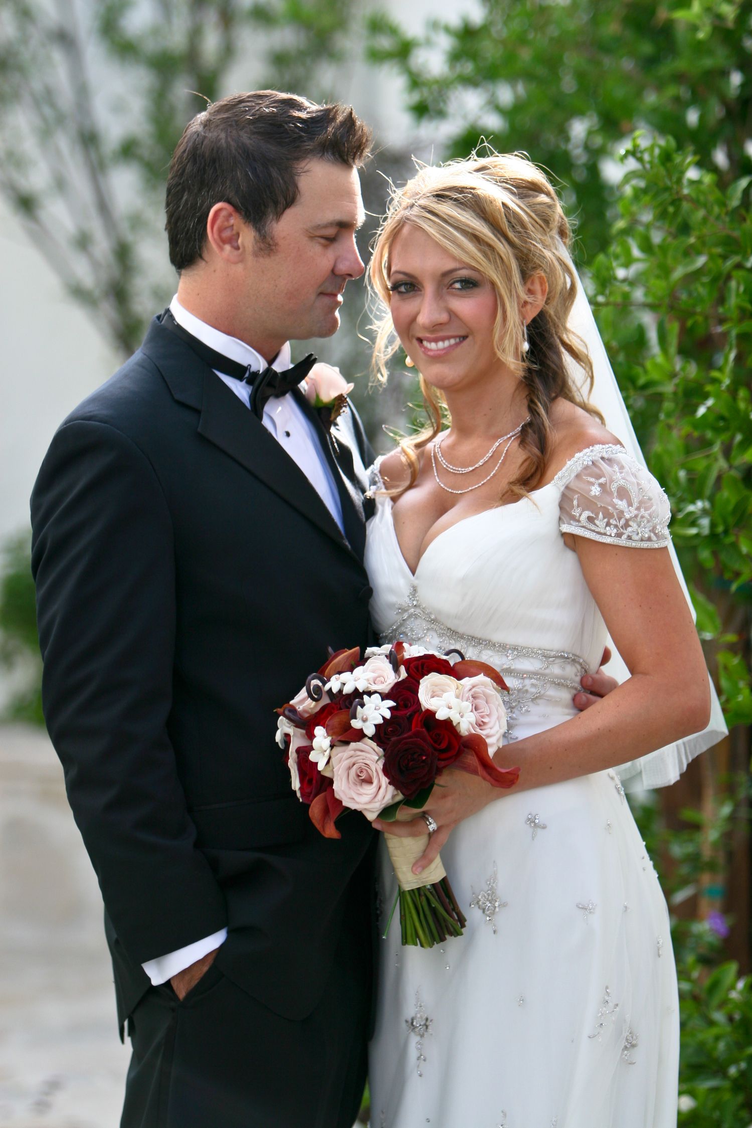 A romantic wedding couple poses with deep red and white floral bouquet in an outdoor garden setting.