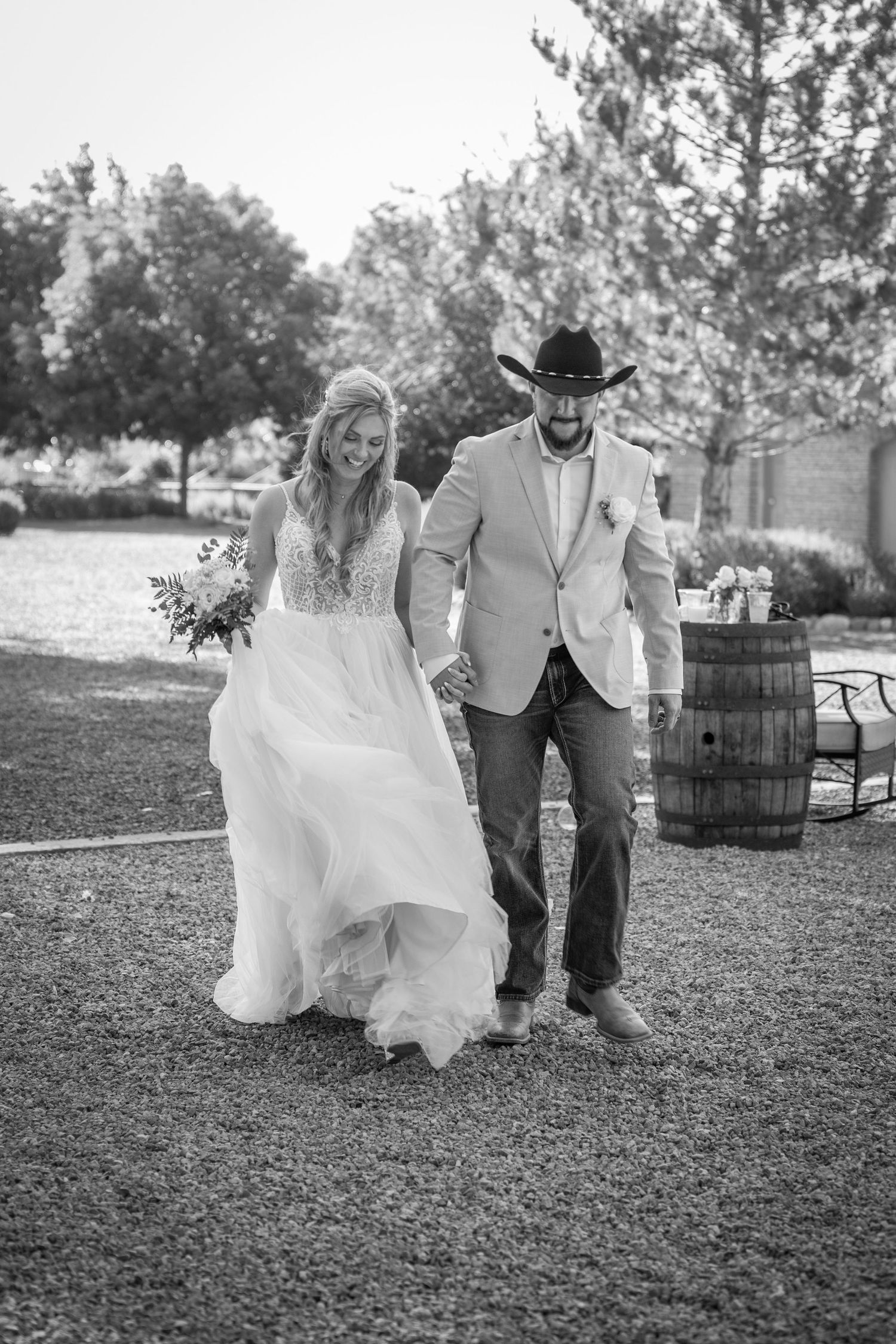 A couple in wedding attire poses playfully on grass near wooden barrels outdoors.