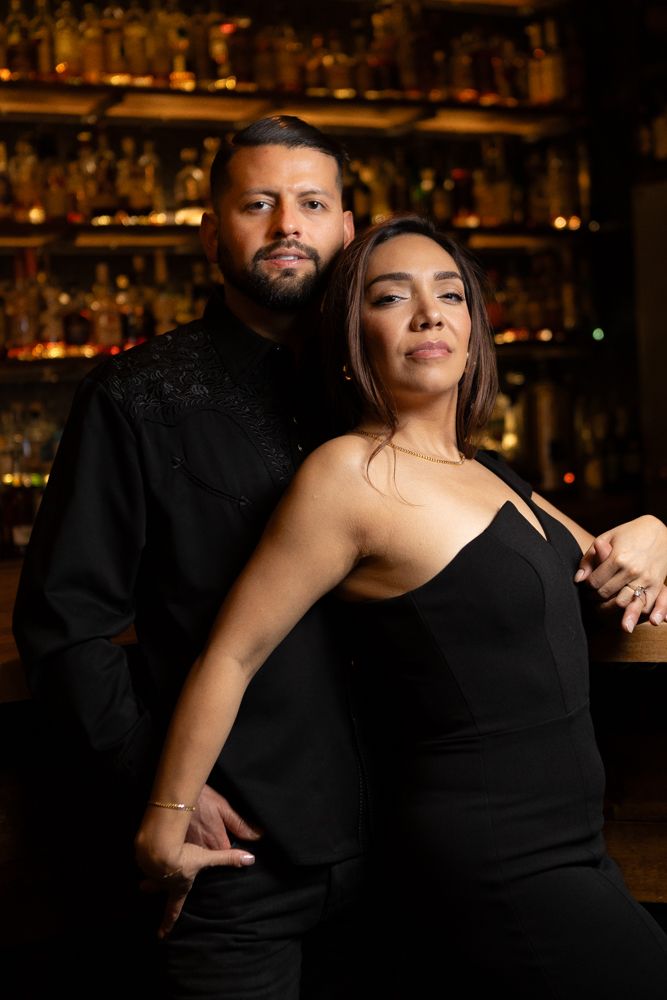 Romantic couple in formal black attire embracing at dimly lit cocktail bar.