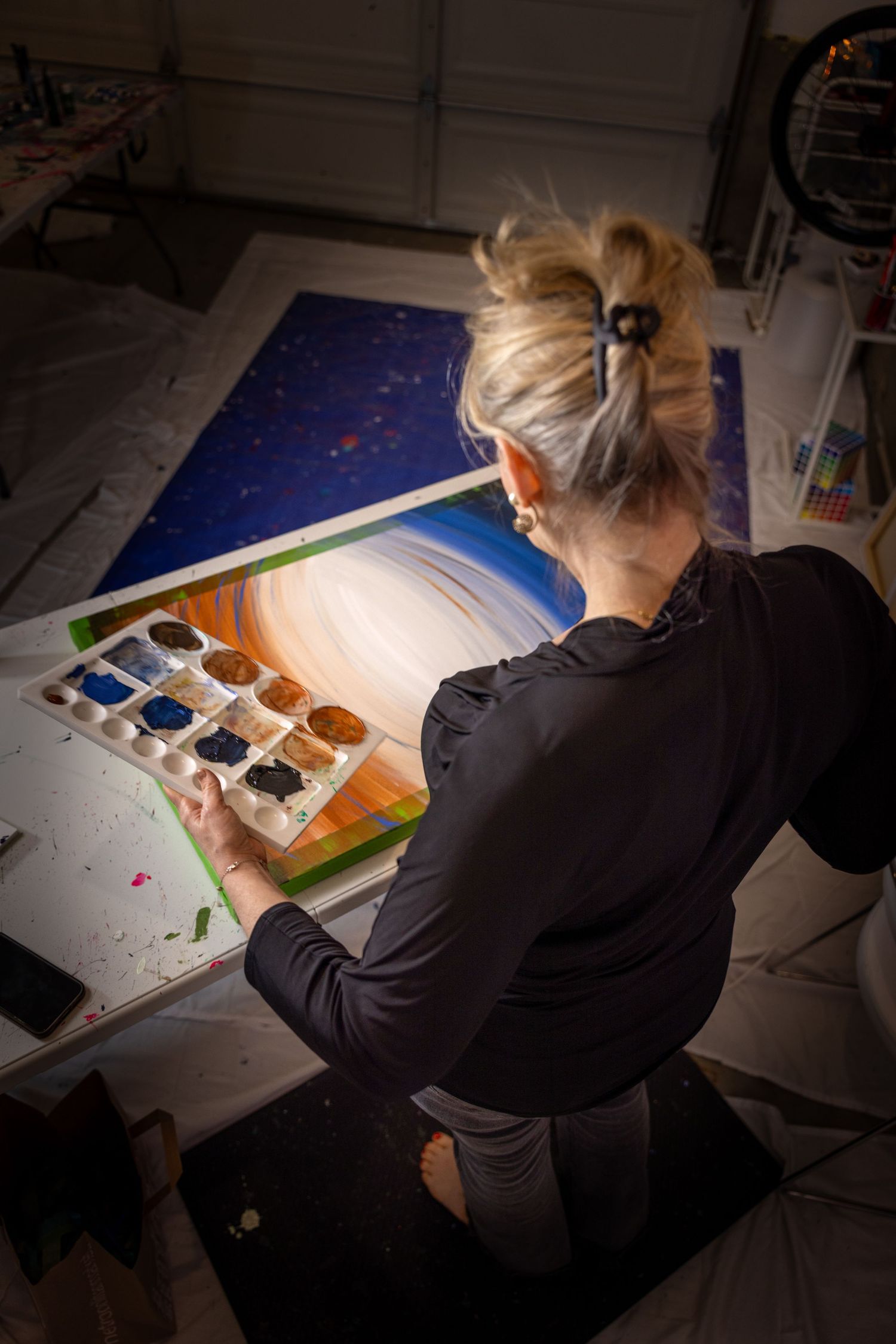 Artist works at desk under focused lighting on detailed watercolor painting project in dark studio setting.