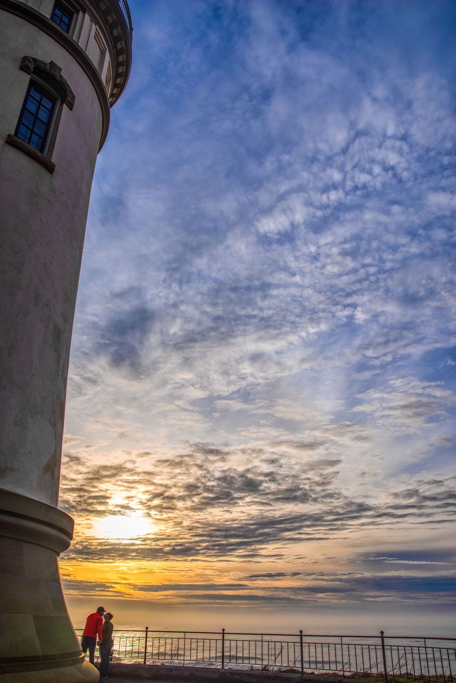 Lifestyle photography of couple in dramatic sunset over ocean horizon viewed from lighthouse observation deck with cloudy sky.