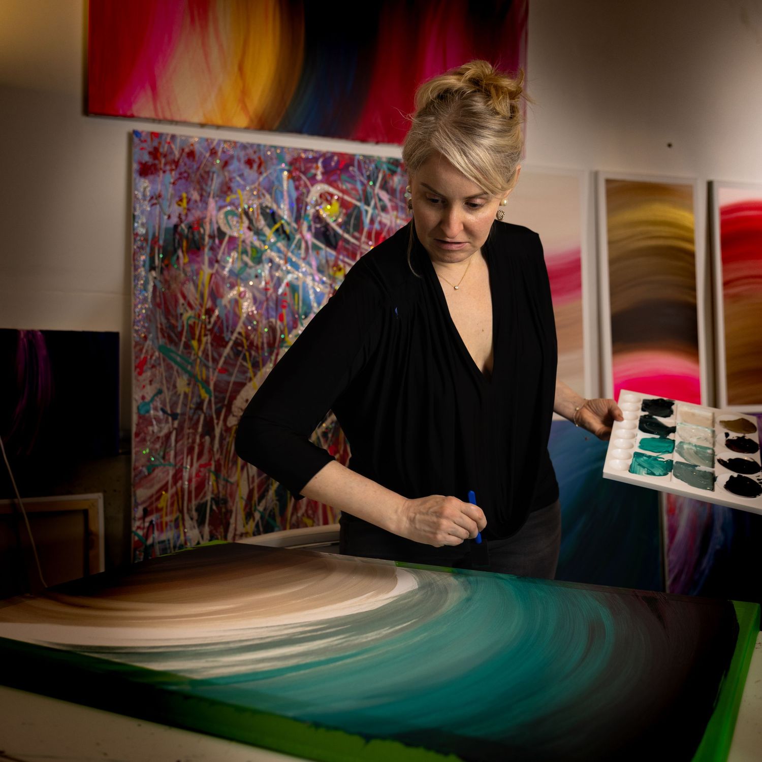 Artist in black attire working on a vibrant teal and white painting in a studio with colorful artwork on display.
