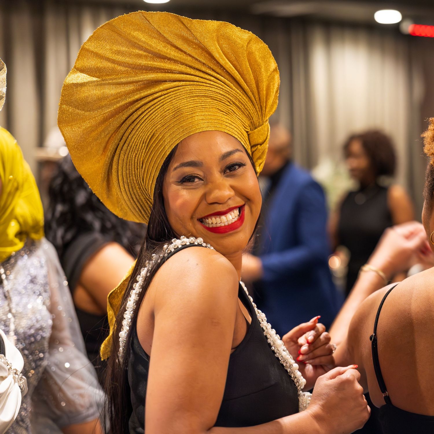 Woman wearing a bright yellow headwrap smiles warmly while seated at a formal event.