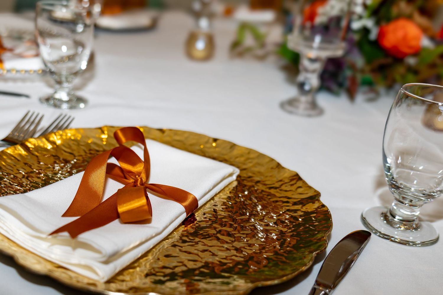 Elegant place setting with gold charger plate, white napkin tied with copper ribbon, and wine glasses on dining table.
