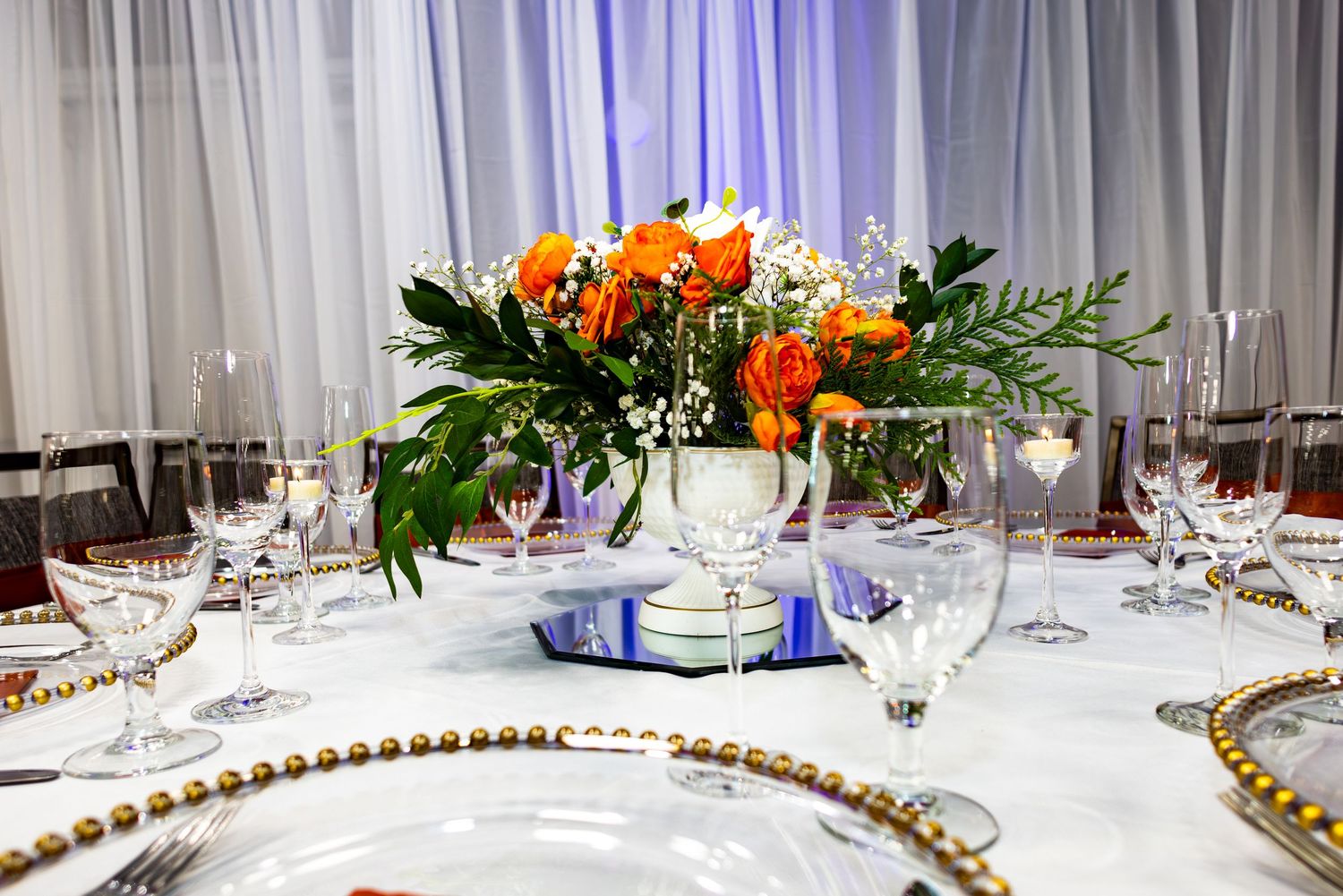 Elegant banquet table settings with white linens, gold beaded chargers, and colorful floral centerpieces in a bright room.