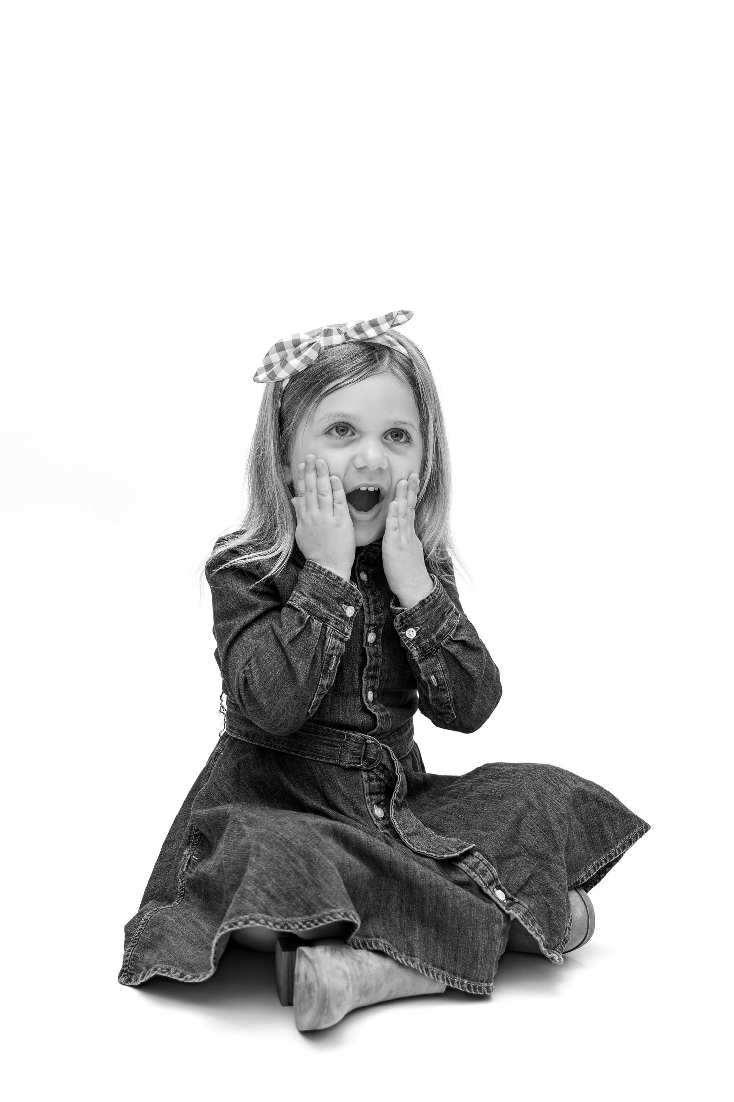 Black and white photo of a surprised little girl in denim dress sitting on floor with hands on cheeks.