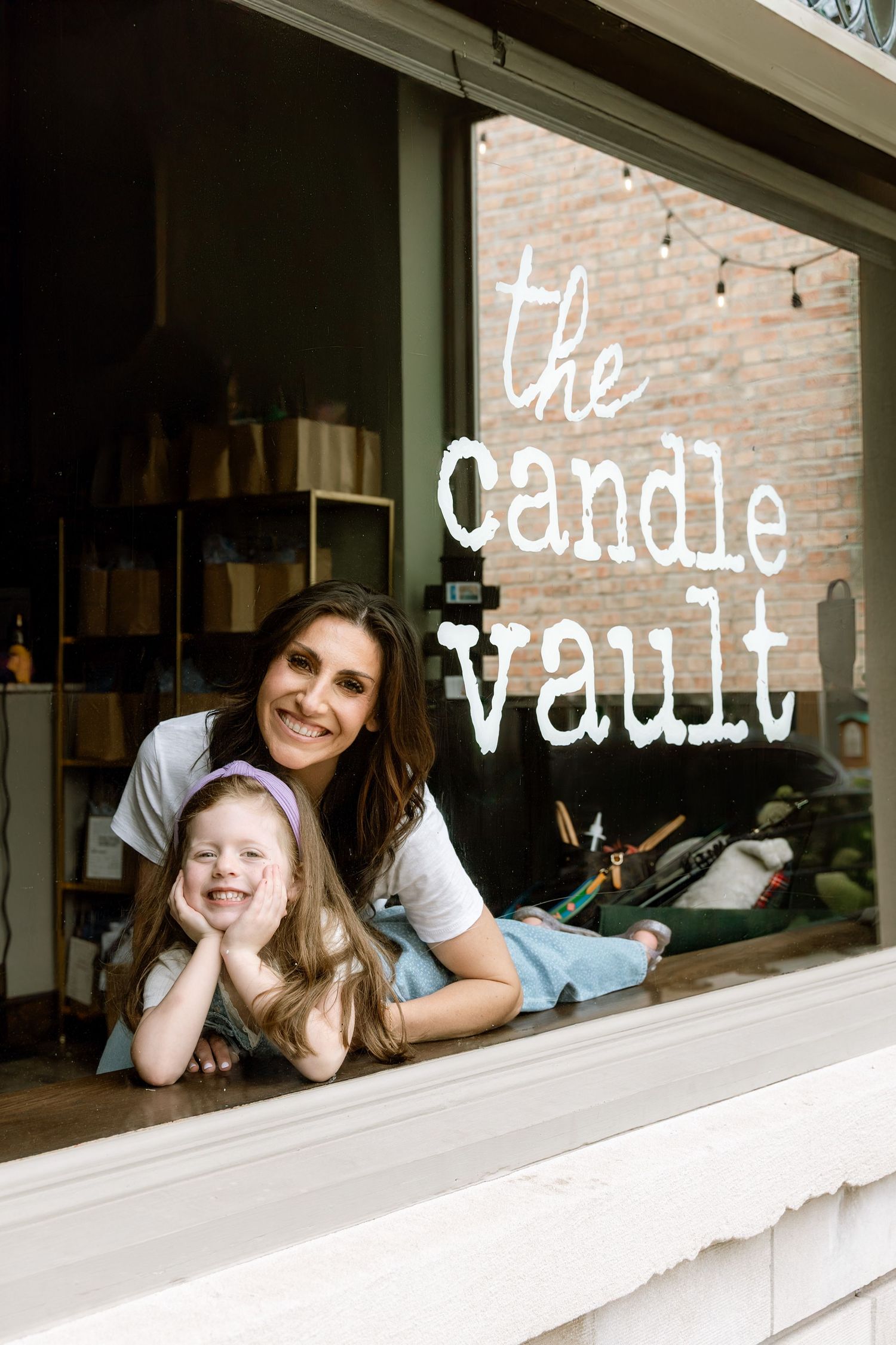 A woman and young girl smile together at the window of The Candle Vault shop.
