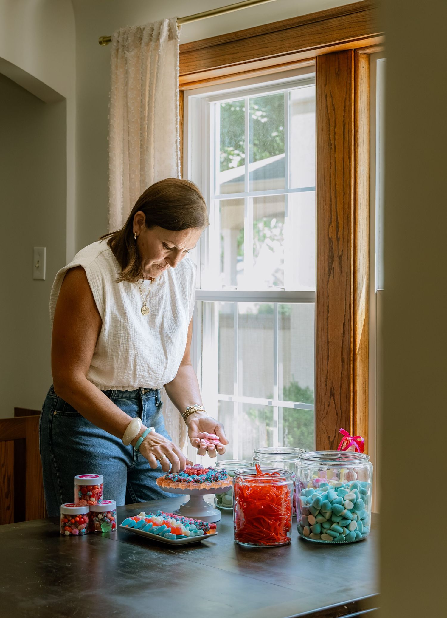 Woman arranging colorful candies in jars on a dark table near a sunlit window in a bright, cozy kitchen.
