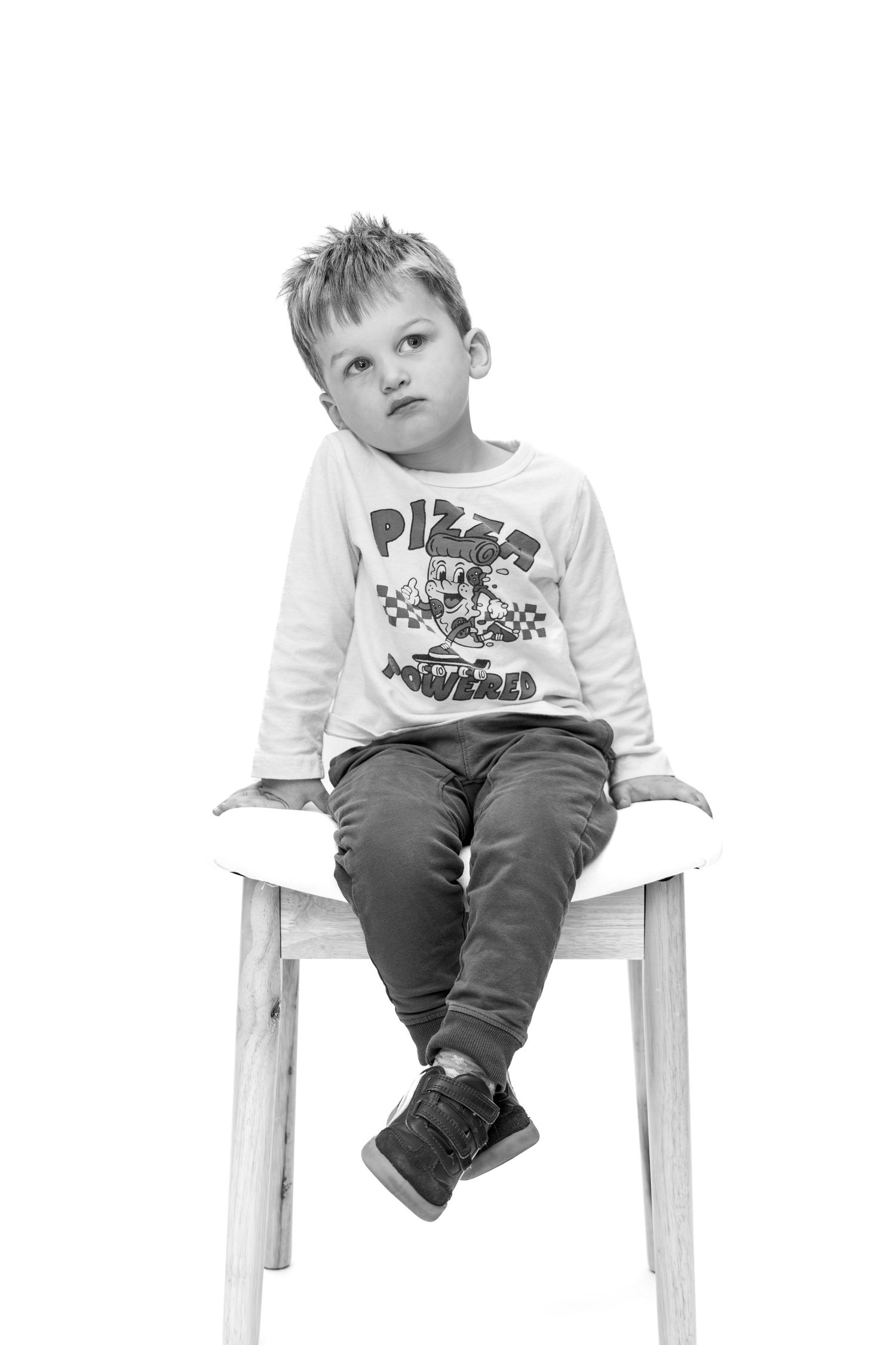 Black and white portrait of a young boy sitting on a wooden chair wearing a Pizza Powered shirt, looking thoughtful.
