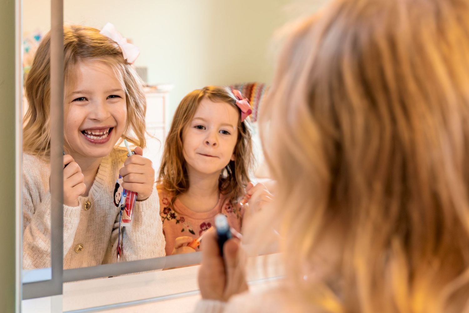 A person with blonde hair smiles while applying makeup and looking in a mirror.