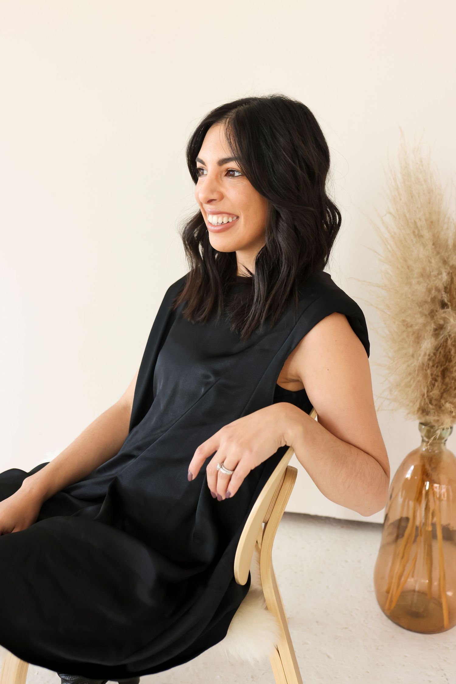 A person in a black sleeveless dress sits in a wooden chair smiling, with pampas grass decor in the background.