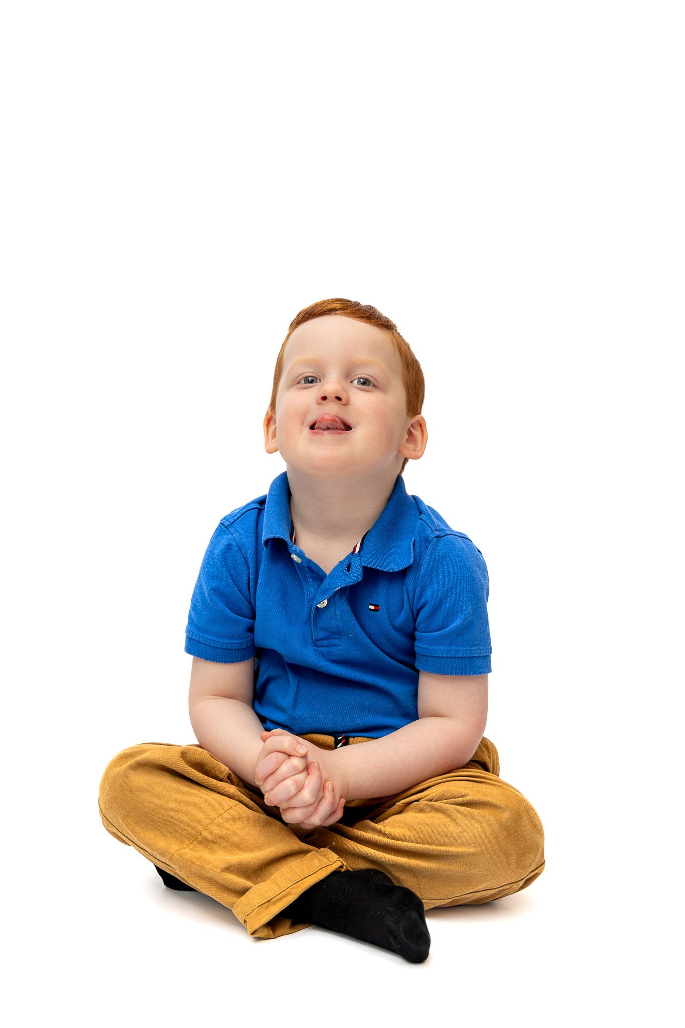 A cheerful red-haired boy in a blue polo shirt and tan pants sits cross-legged on a white background, looking up.
