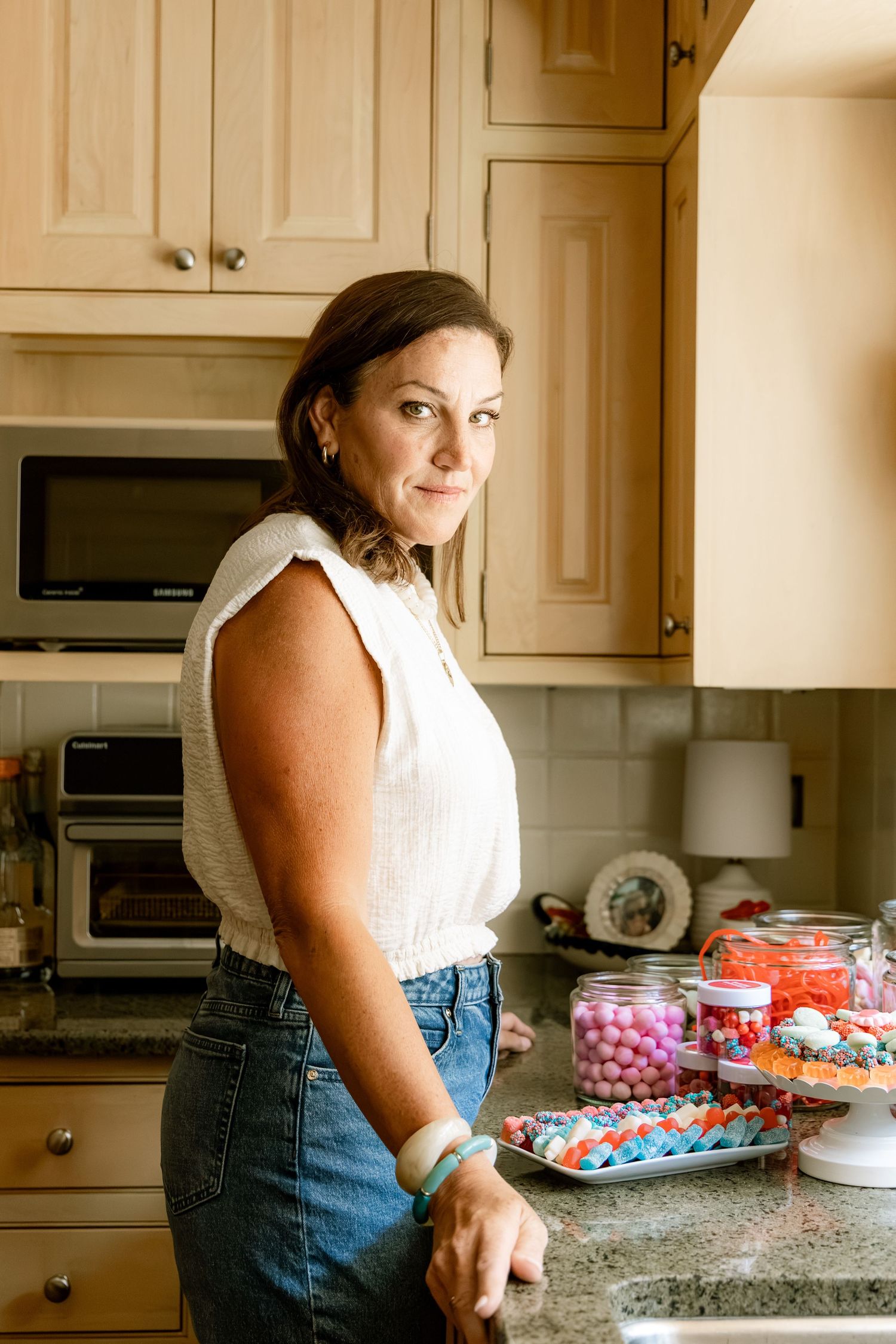 Woman in white top and jeans arranging colorful candies on kitchen counter with jars of sweets nearby.