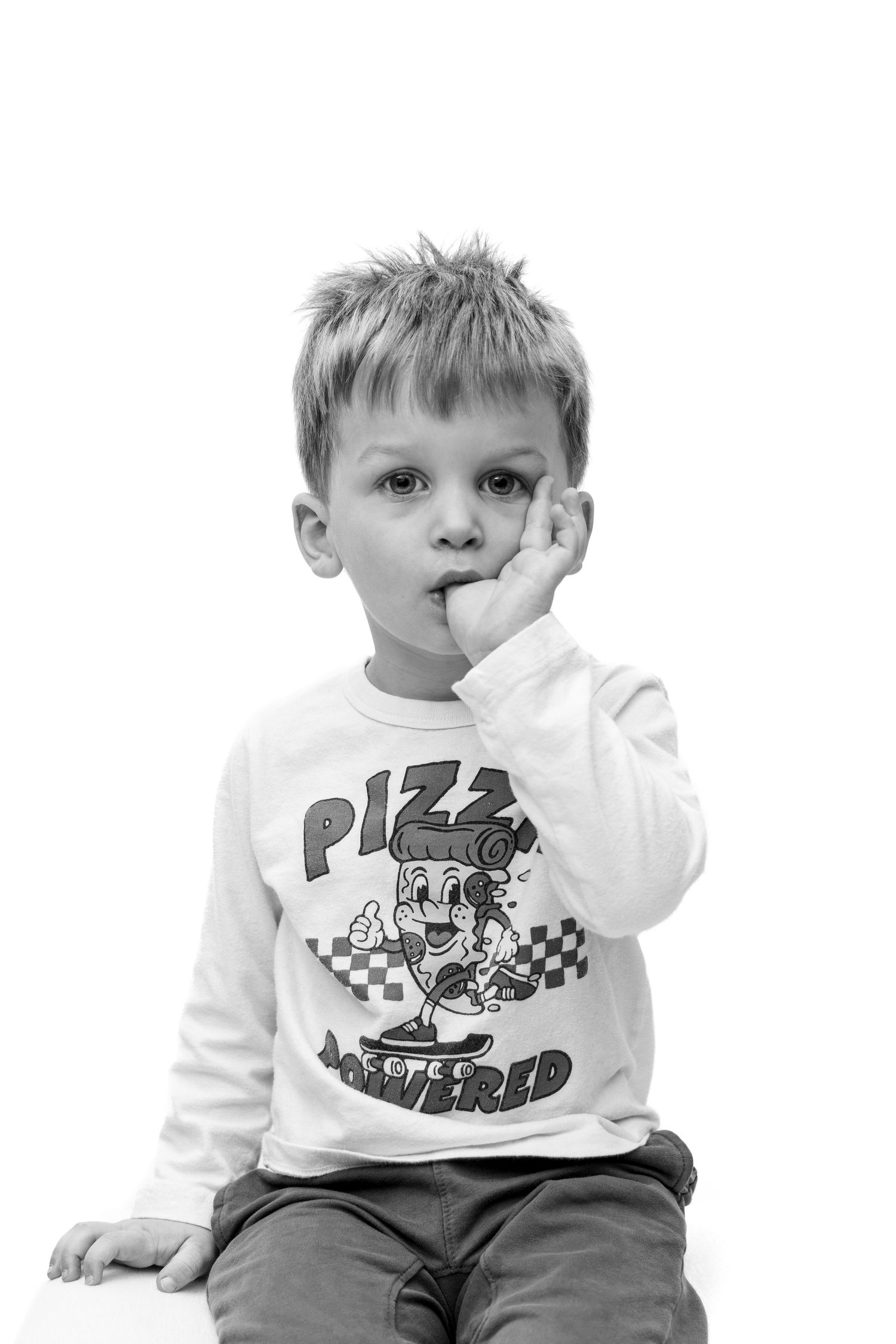 Black and white photo of a young boy in a pizza-themed shirt sitting thoughtfully with his hand near his mouth.