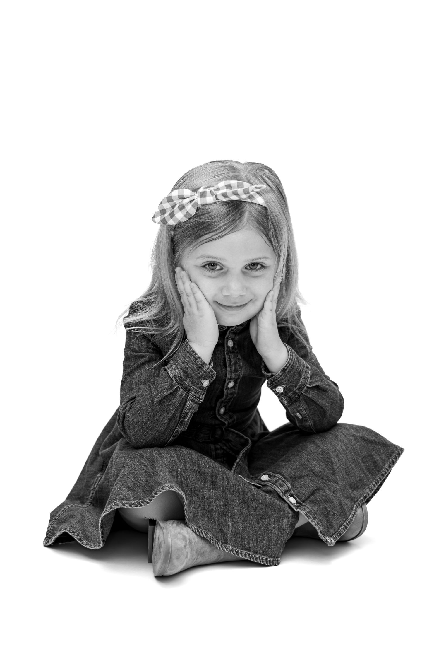Black and white photo of a cute young girl sitting cross-legged, hands on cheeks, wearing denim outfit and bow headband.