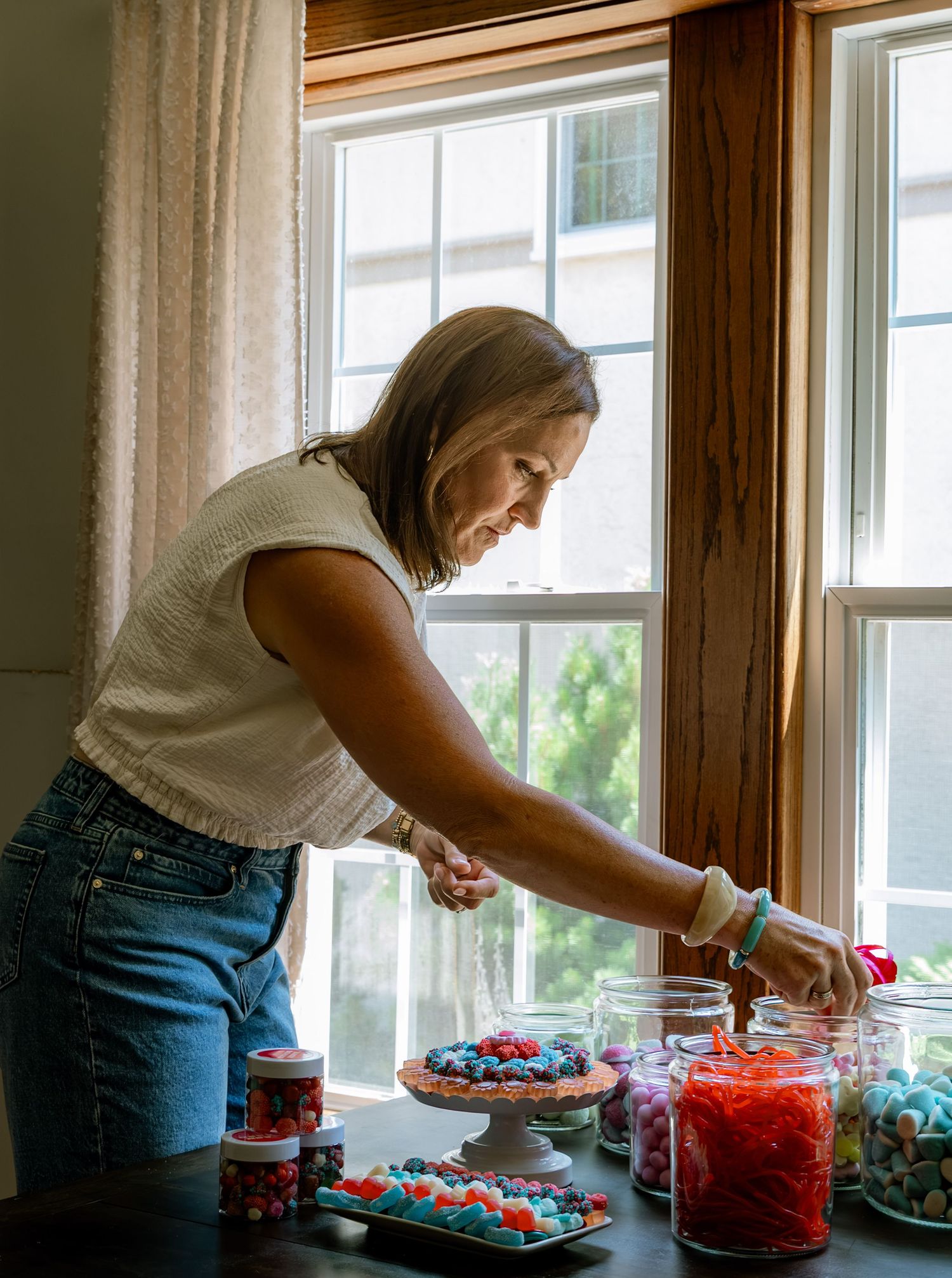 Woman arranging colorful candy display on dark table near window with jars of sweets and treats.