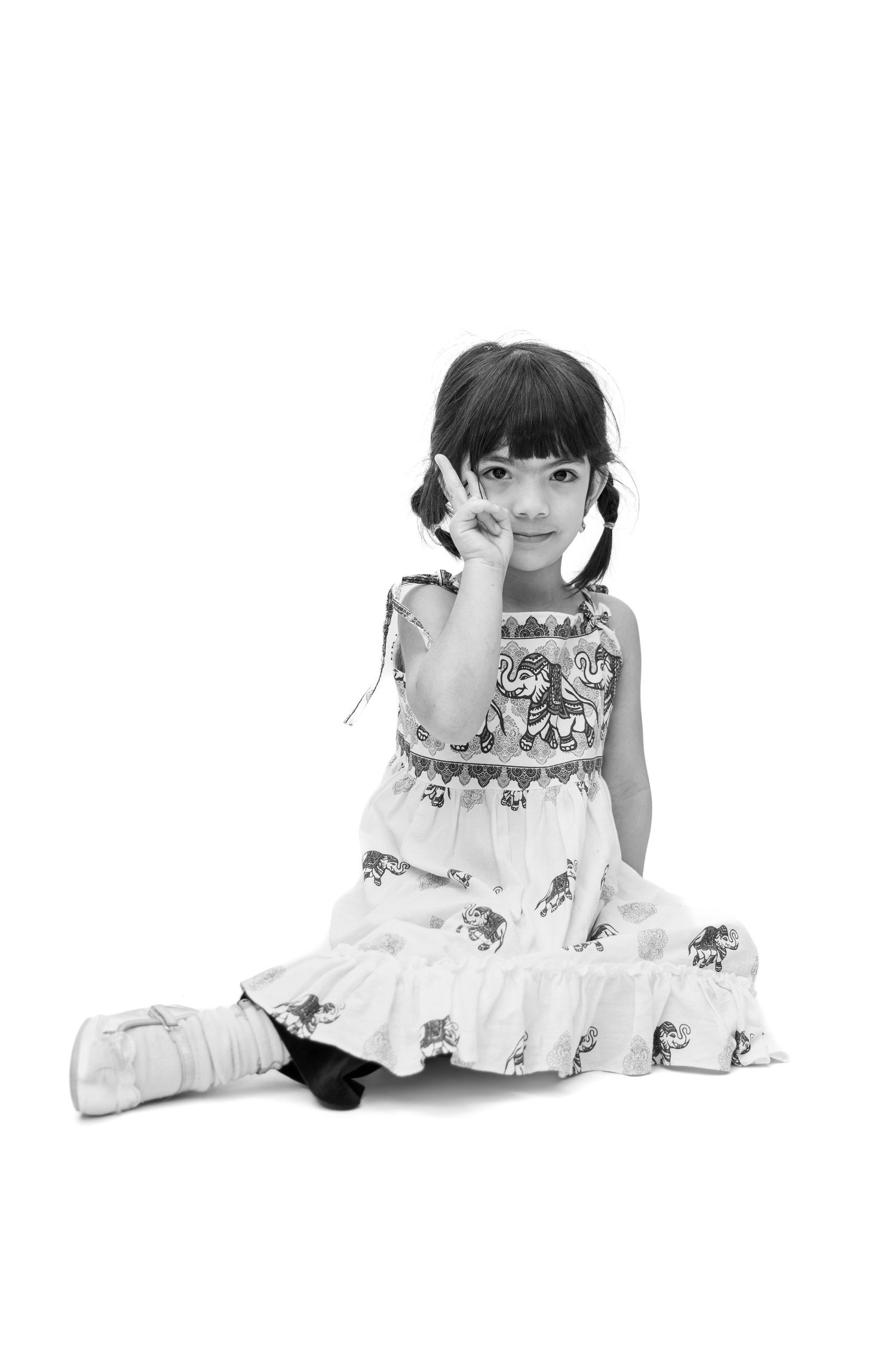 Black and white photo of a young Asian girl in a patterned dress sitting on floor, making a peace sign gesture.