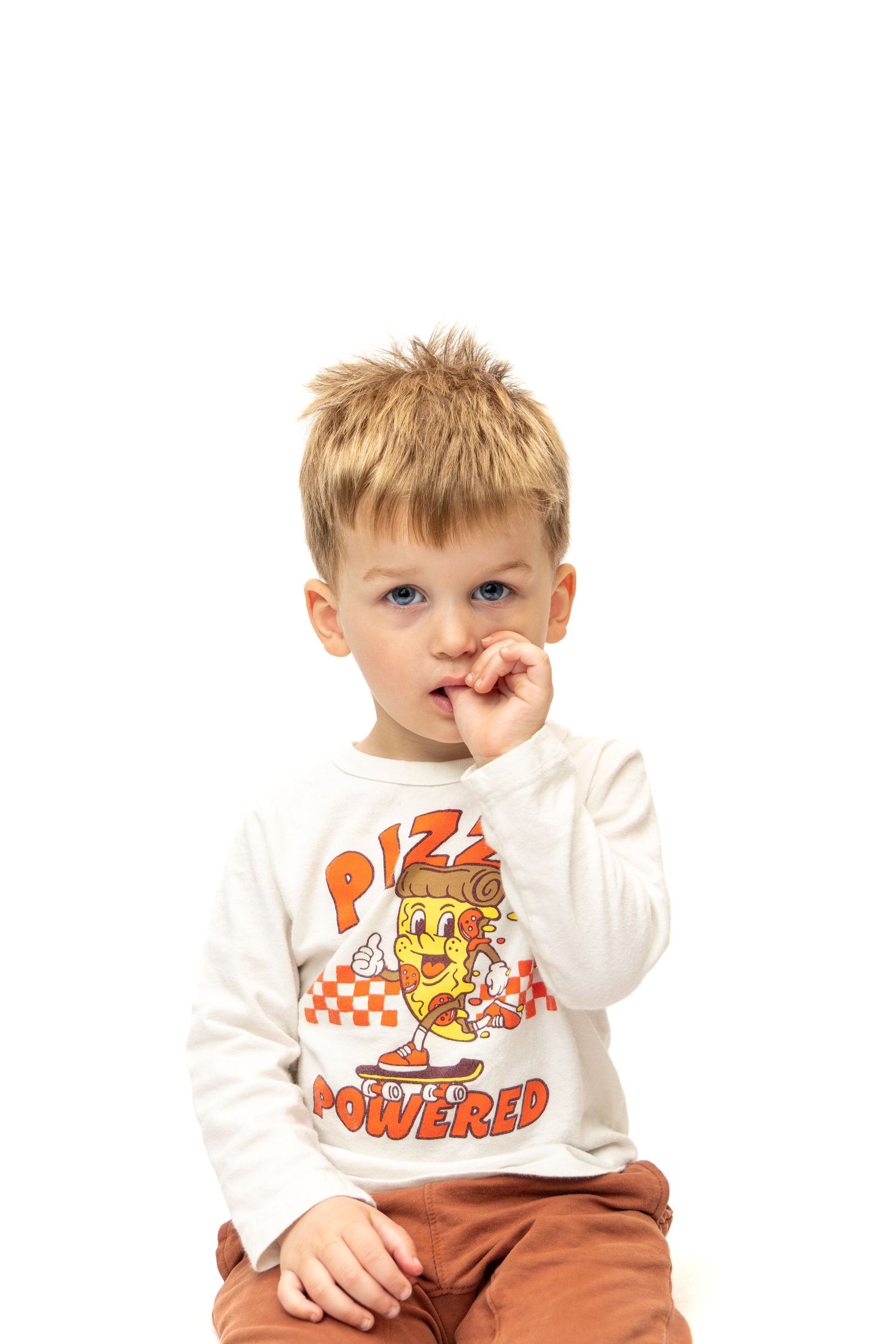 A young blonde boy with blue eyes wearing a white pizza-themed long sleeve shirt and brown pants.