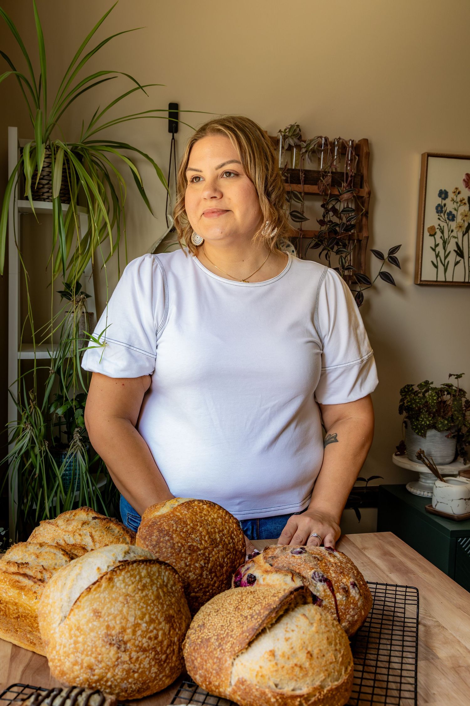 Woman in white t-shirt stands proudly behind a cooling rack of freshly baked artisan sourdough breads in a cozy home kitchen.