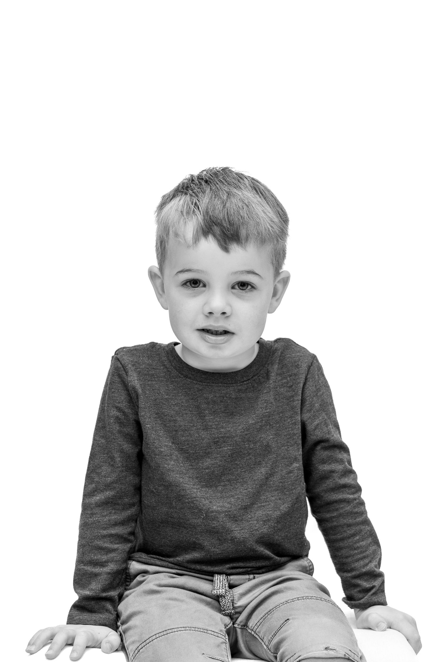 Black and white portrait of a young boy in a long-sleeve shirt and jeans sitting against a white background.