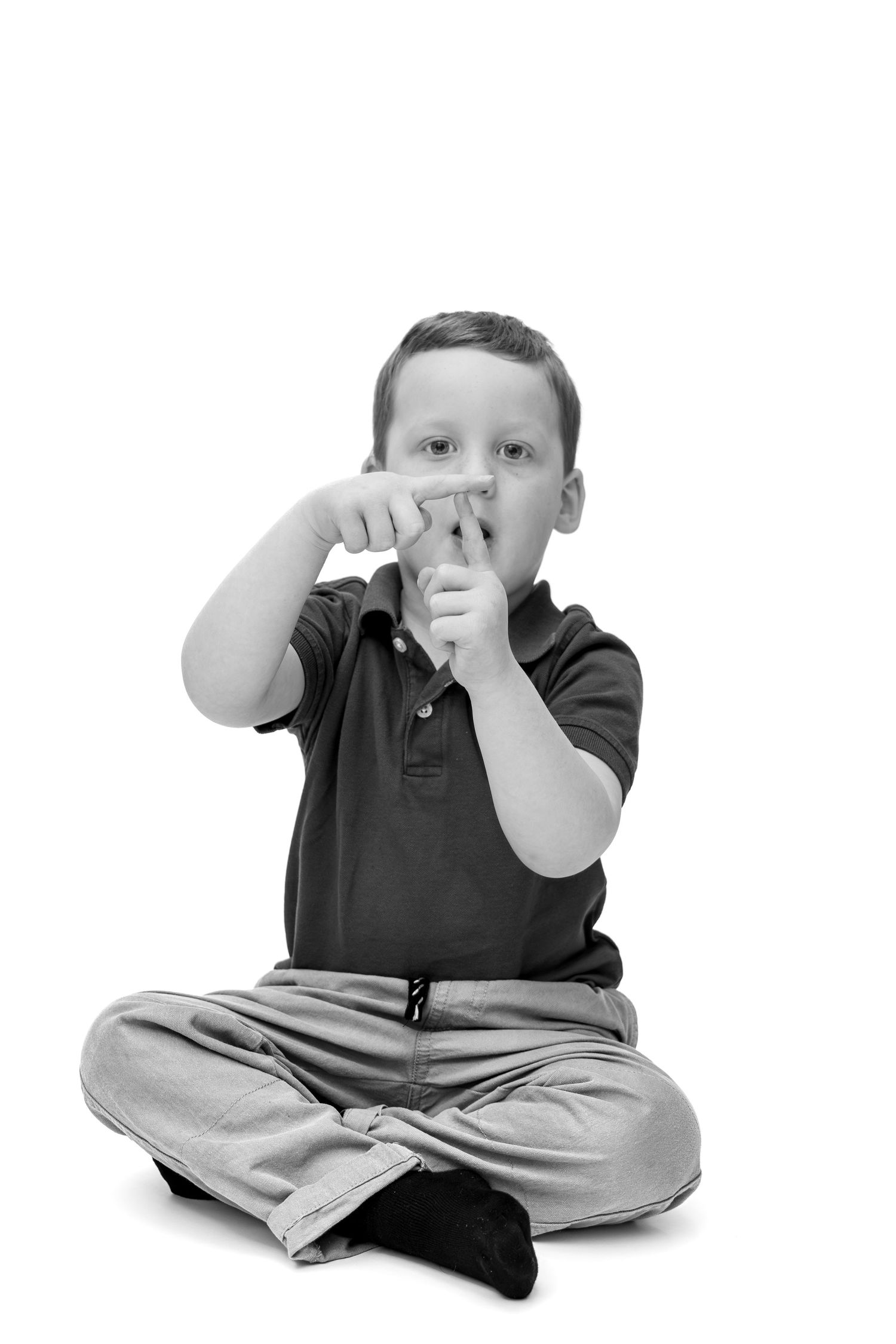 Black and white photo of a young boy sitting cross-legged, pointing fingers forward toward the camera.