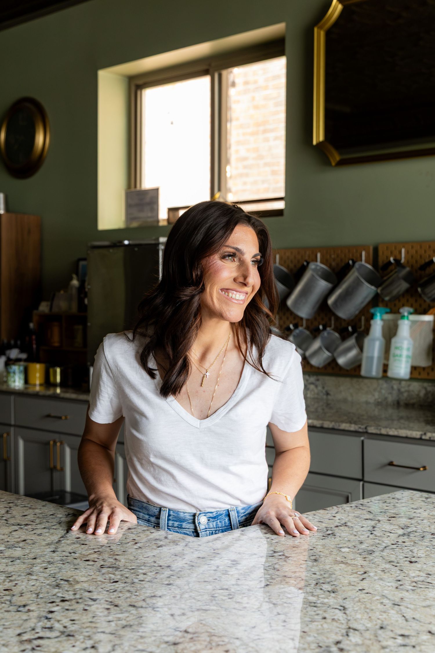 Smiling brunette woman in white v-neck top and jeans leaning on granite kitchen counter with green walls.