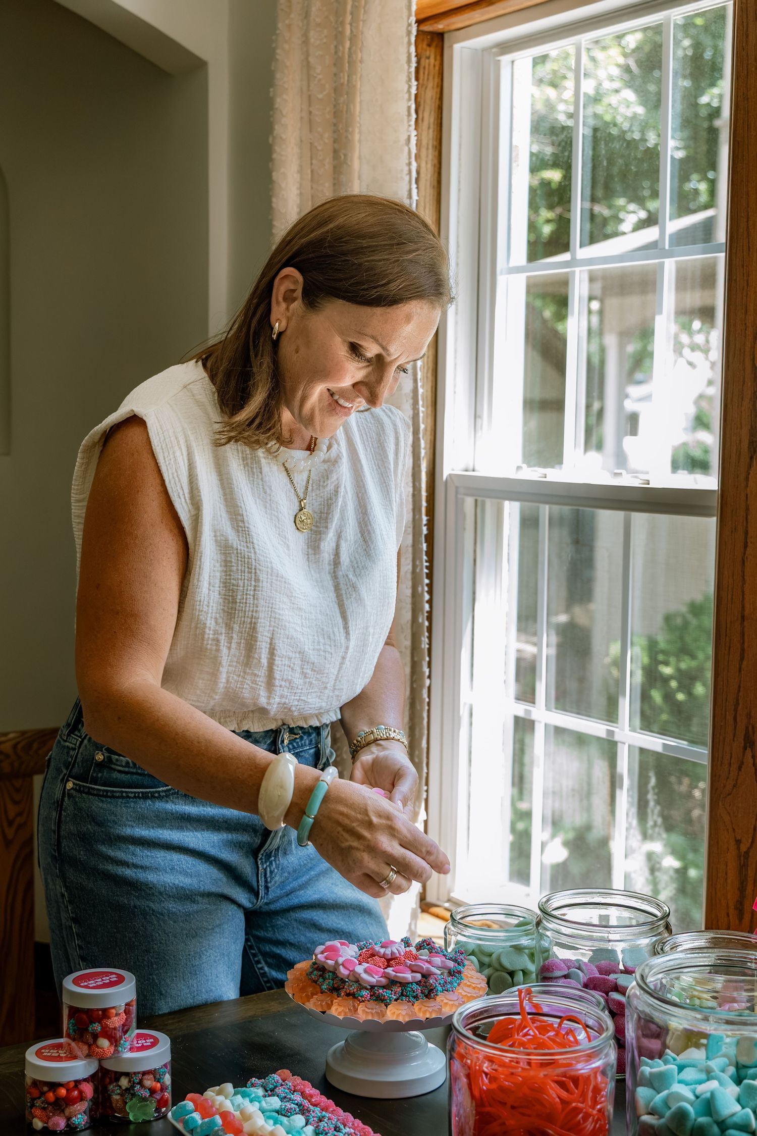 Woman decorating a colorful celebration cake surrounded by jars of candy near a sunlit window.