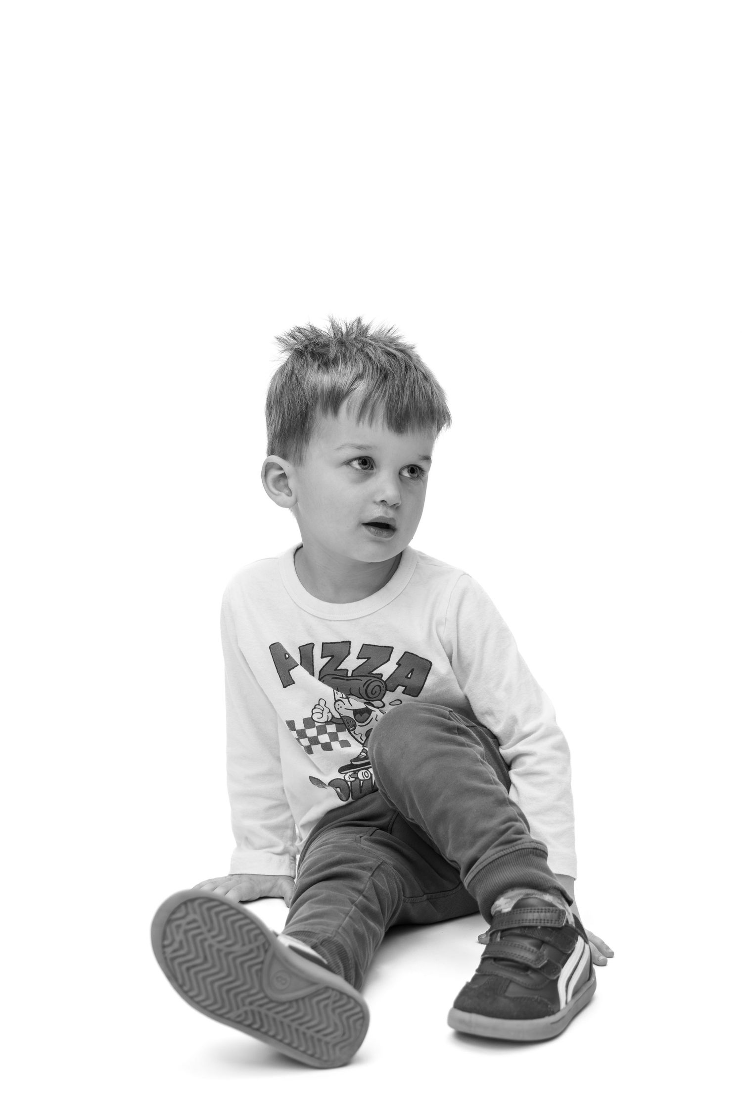 Black and white photo of a young boy sitting on floor wearing a pizza shirt, jeans, and sneakers, looking sideways.