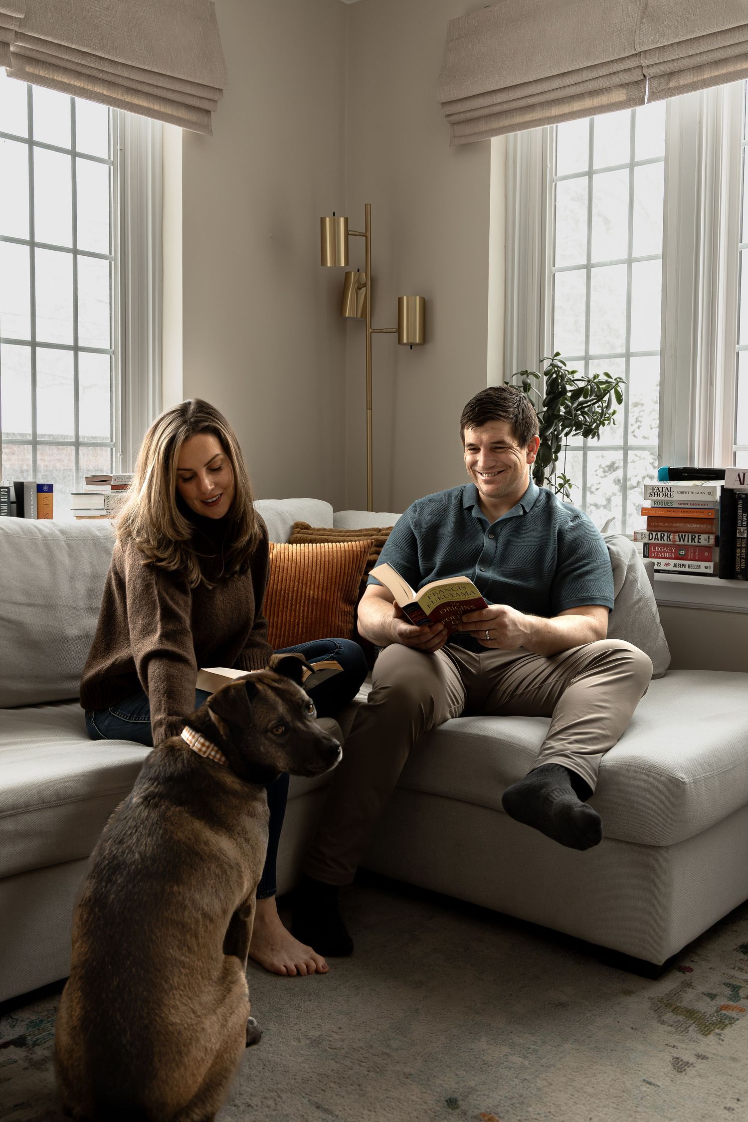 A couple relaxes on a couch while reading books together in a bright, window-lit room.