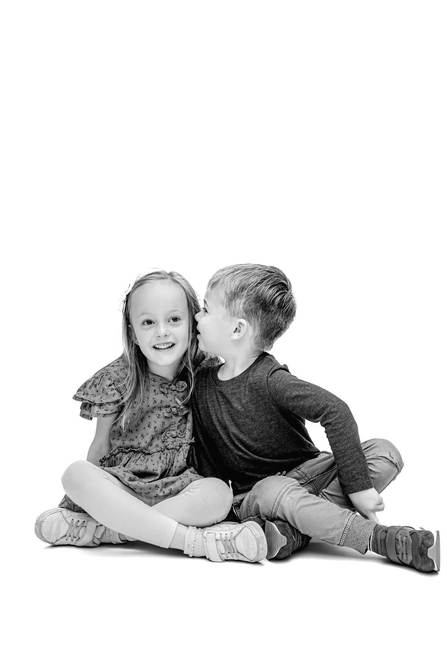 Black and white photo of two young children sitting on floor, boy whispering in smiling girls ear.
