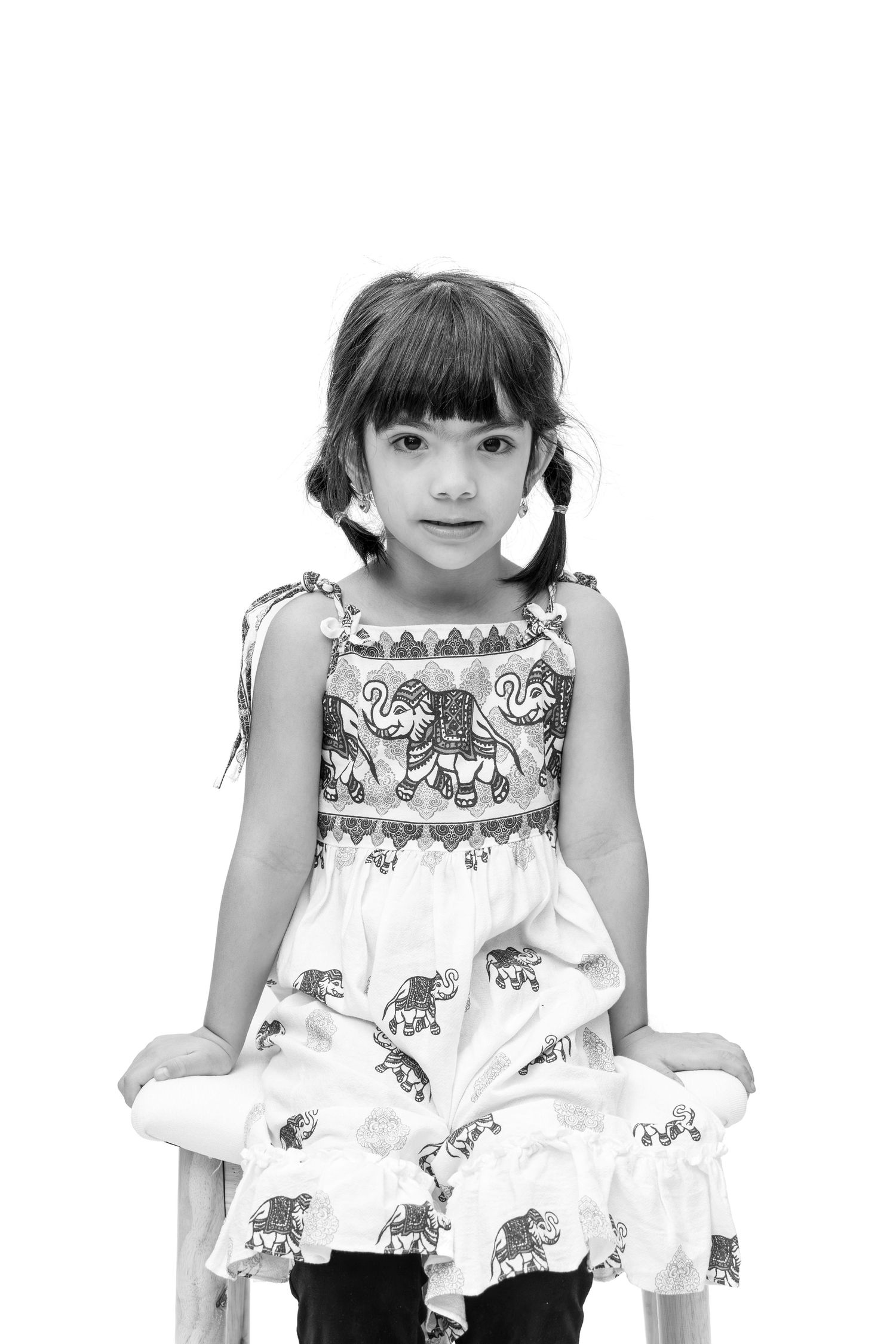 Black and white portrait of a young girl in an elephant print dress with pigtails, sitting on a chair against white background.