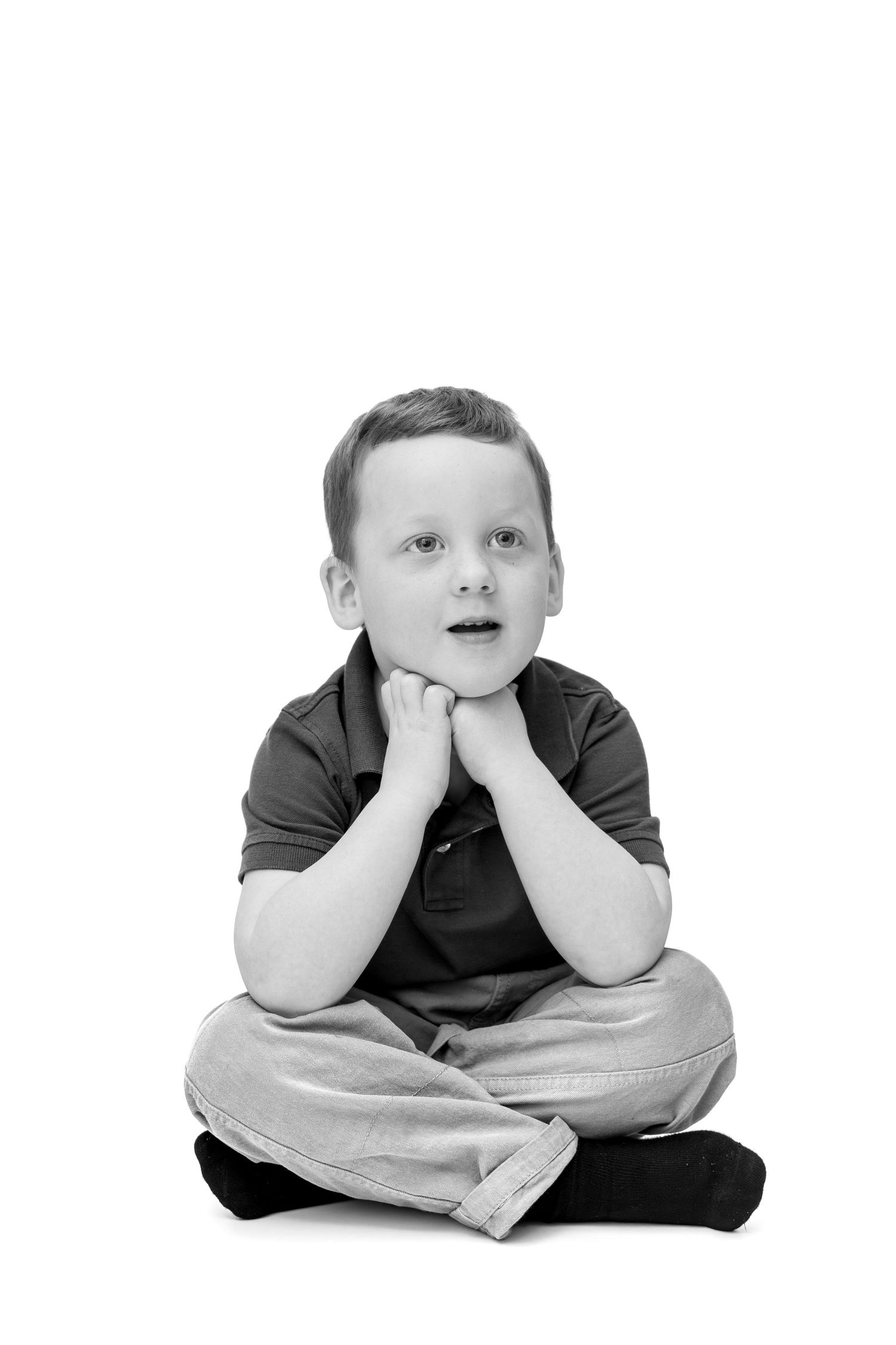 Black and white photo of a young boy sitting cross-legged with hands on chin, looking up with a curious expression.