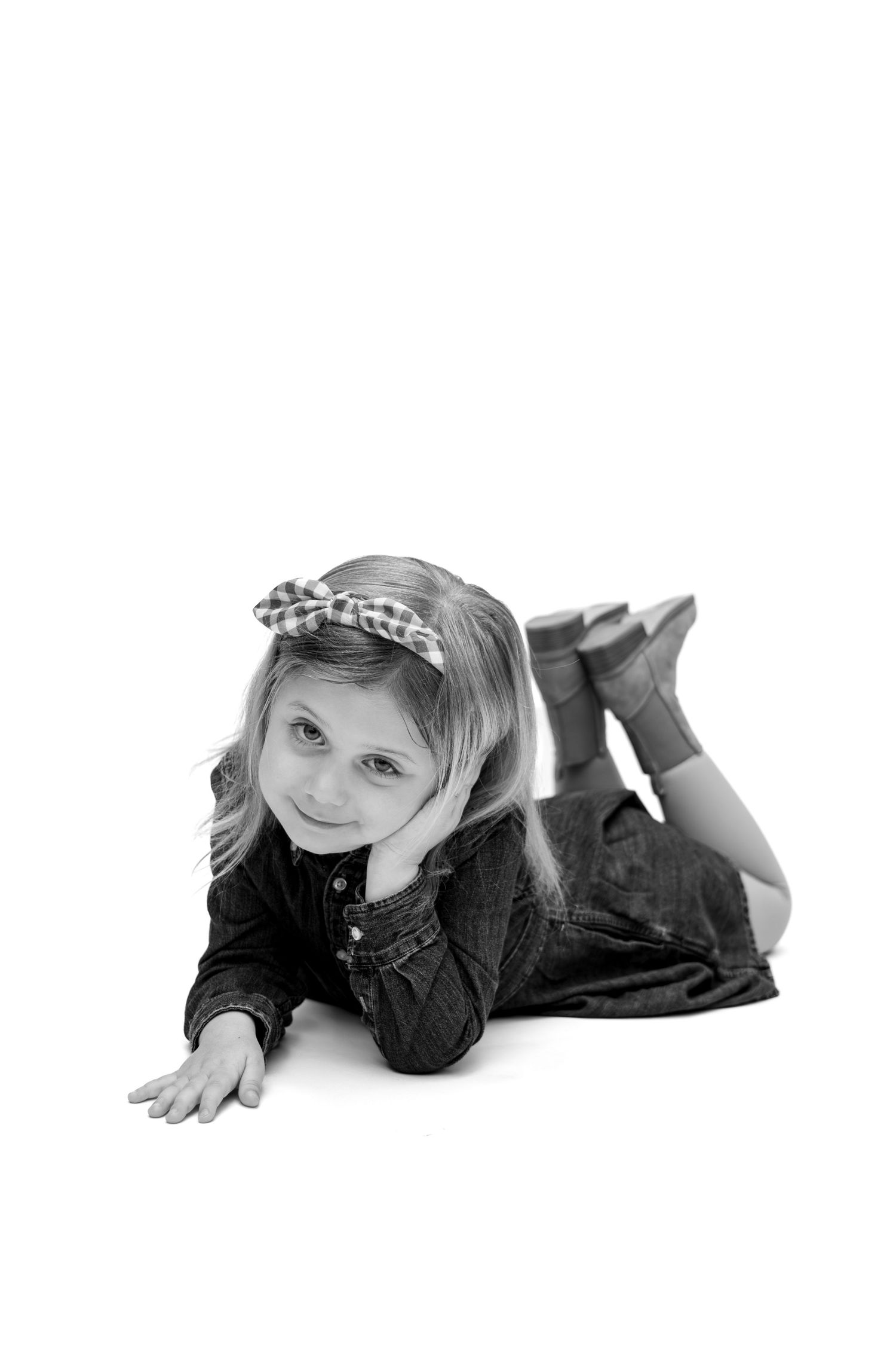 Black and white photo of a cute young girl lying on floor, wearing denim dress and leopard print headband.