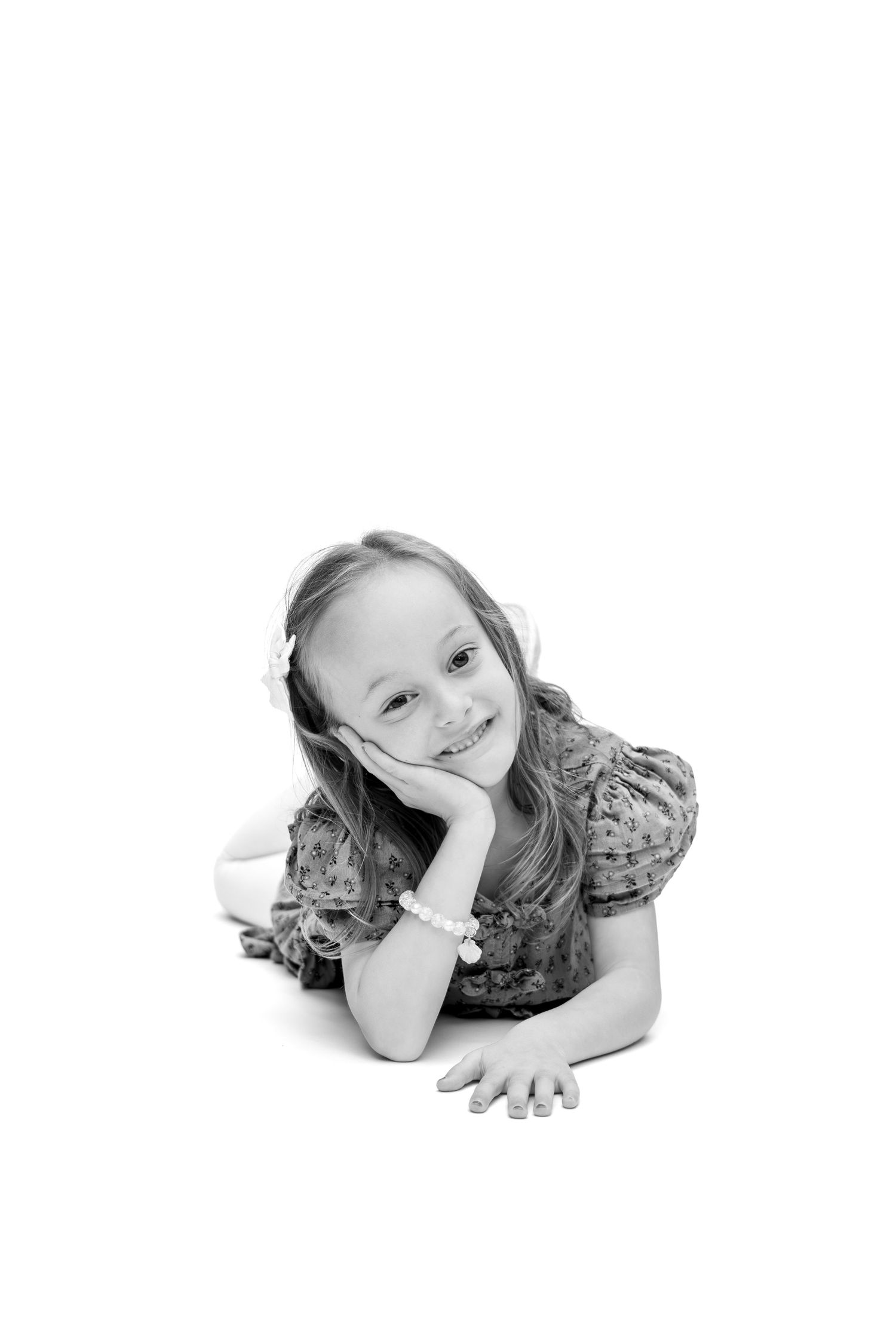 Black and white photo of a smiling young girl lying down, resting her chin on her hand, wearing a floral dress.