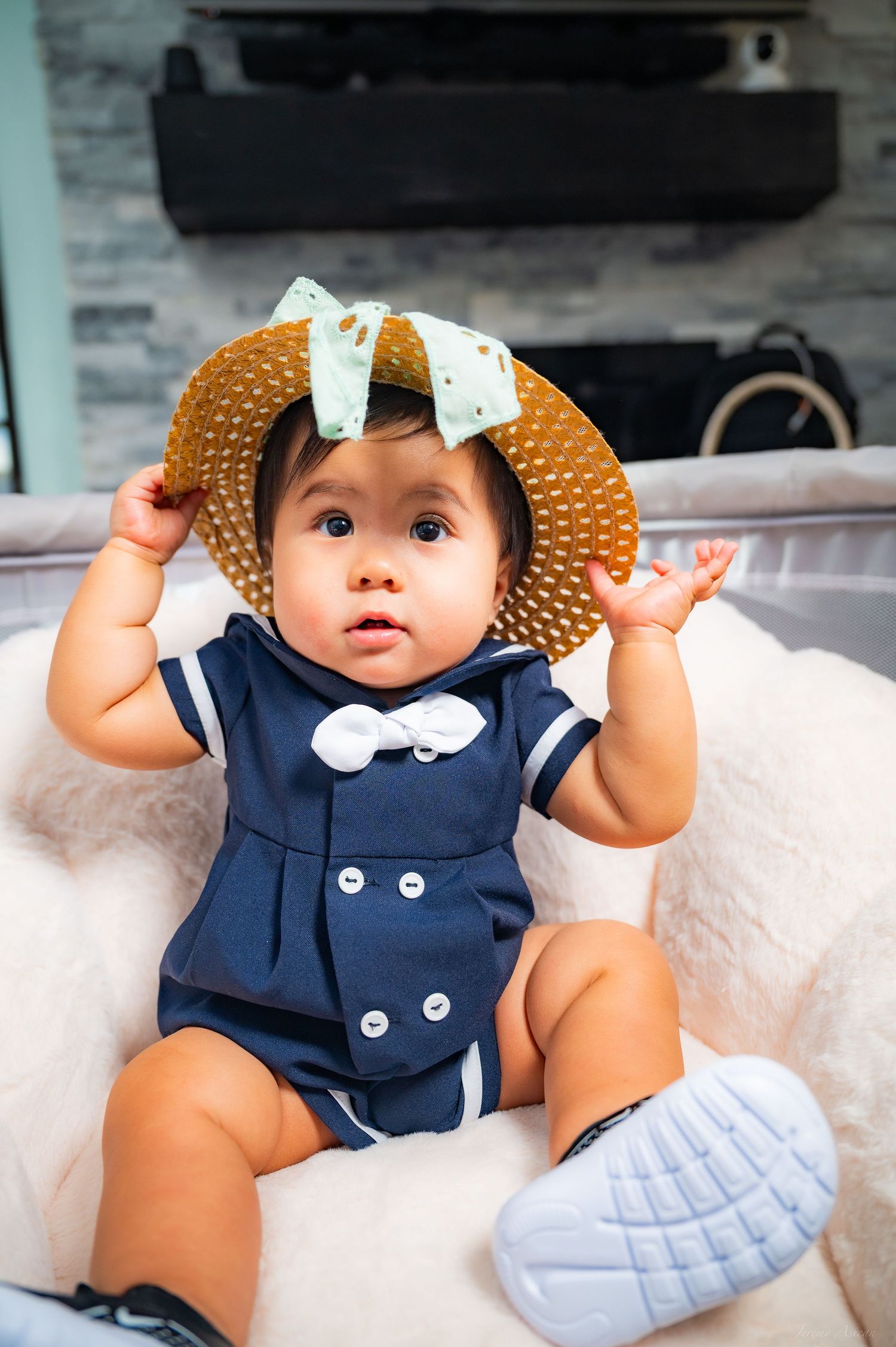Adorable baby in navy sailor romper and straw hat with bow sits in fluffy white chair, holding hat with tiny hands. Stockton photographer