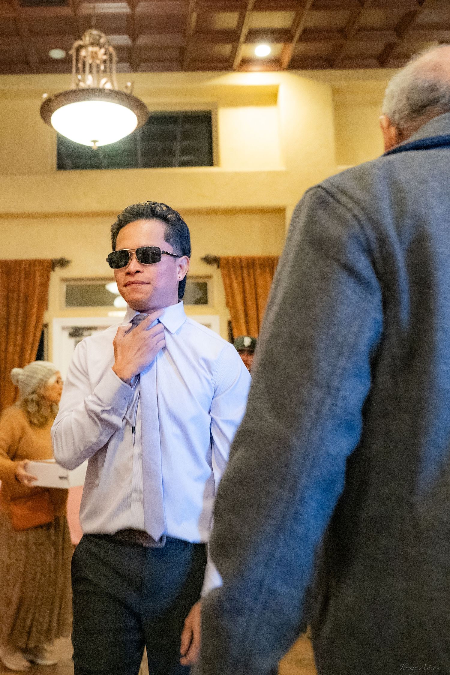 Young man in sunglasses adjusting his tie at a formal indoor event, wearing a light blue dress shirt.