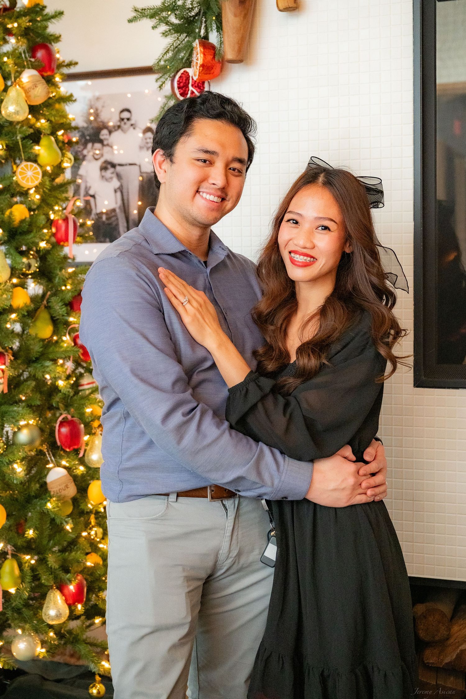 A smiling Asian couple poses together near a decorated Christmas tree, man in blue shirt and woman in black dress. Stockton photographer