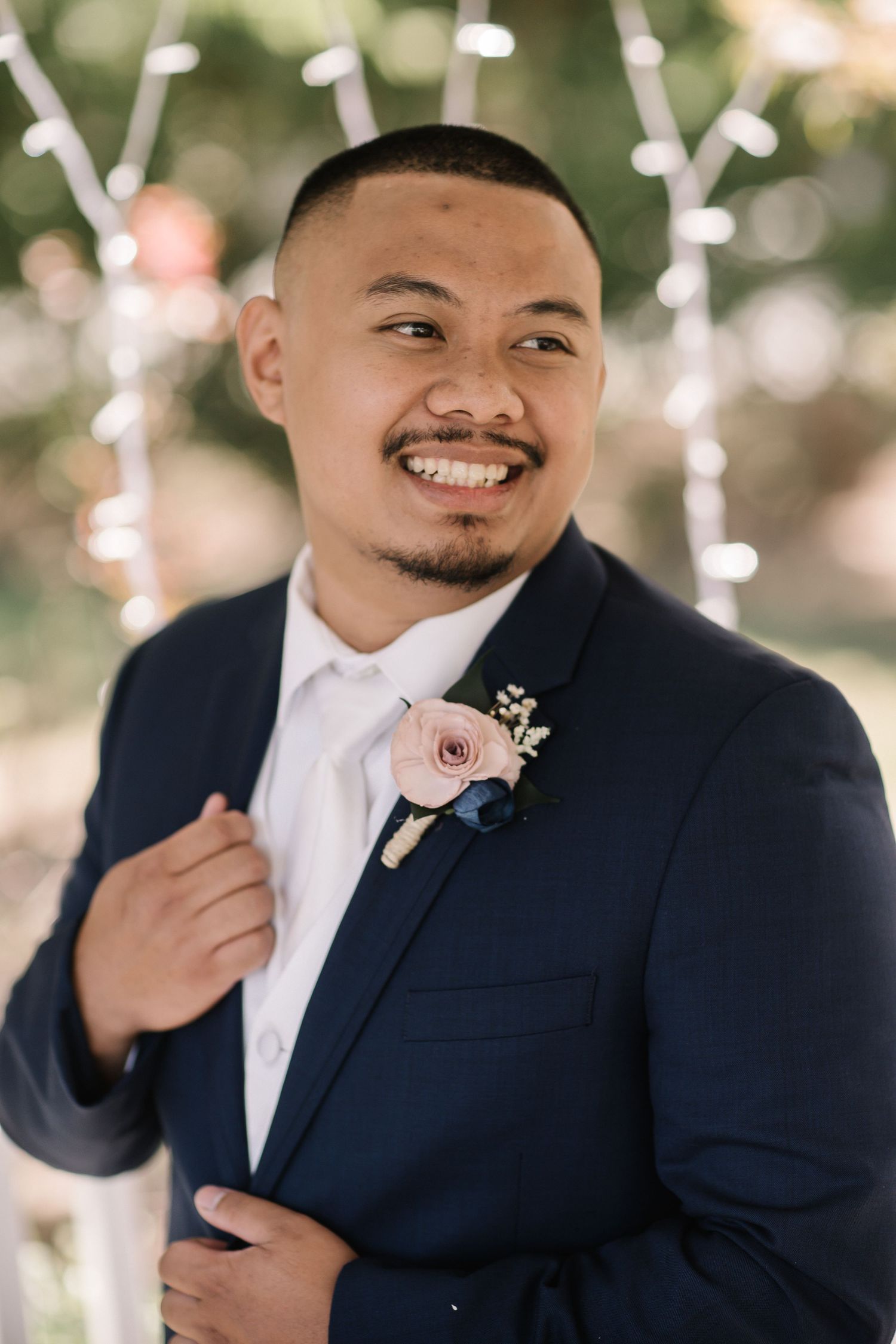 Smiling groom in navy suit with pink boutonniere at outdoor wedding ceremony with bokeh lights. Stockton photographer