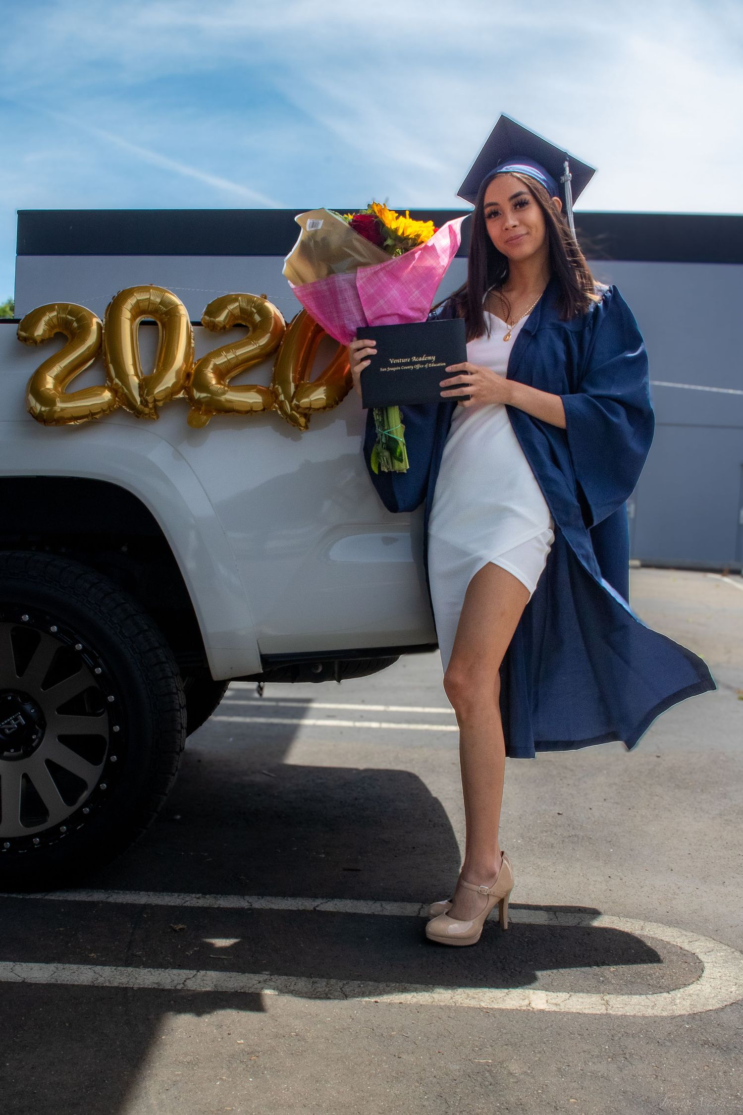 Young woman in navy graduation cap and gown holding diploma beside white truck with 2020 balloons and flowers.
