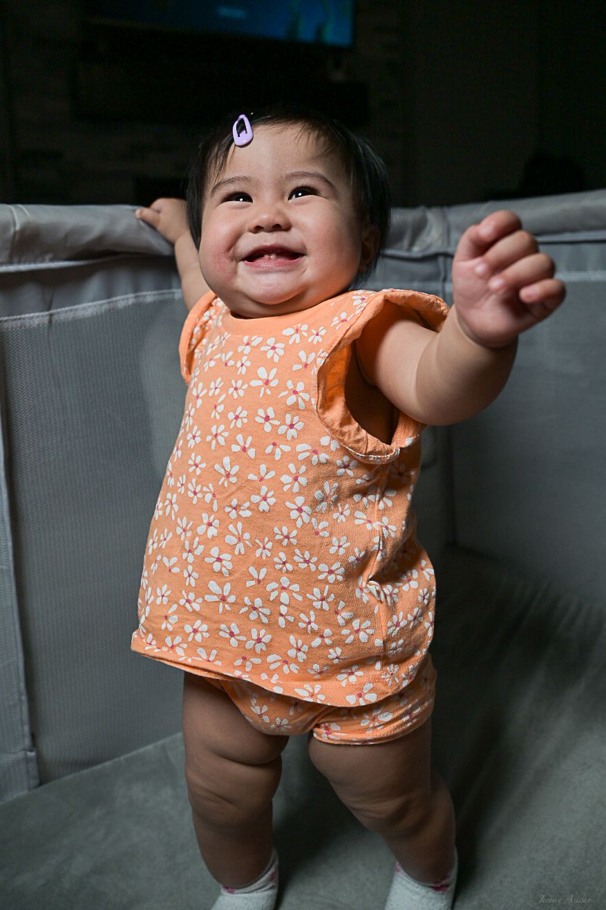 Smiling baby girl in orange floral outfit standing against a gray sofa, reaching out with one hand.