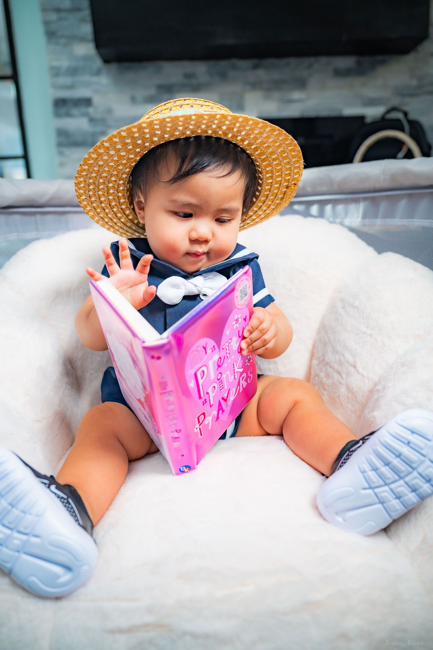 Cute baby in straw hat reading a pink book while sitting in a fluffy white chair wearing blue sneakers. Stockton photographer