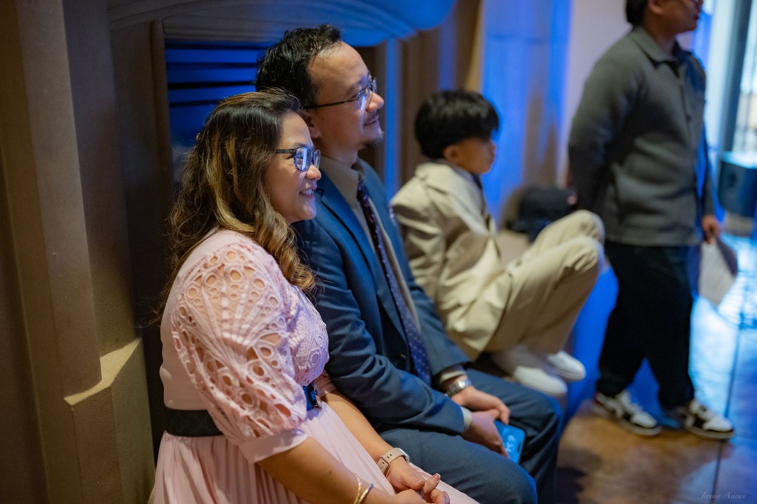 A couple sits together against a wooden wall, smiling, in a softly lit blue ambiance at an indoor event.