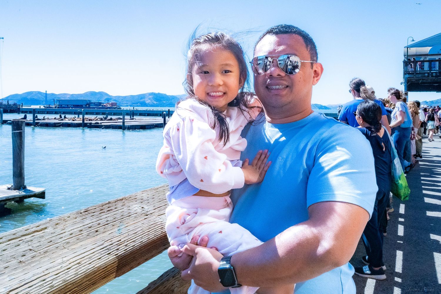 A man holding a young girl near a waterfront pier on a sunny day, smiling at the camera.