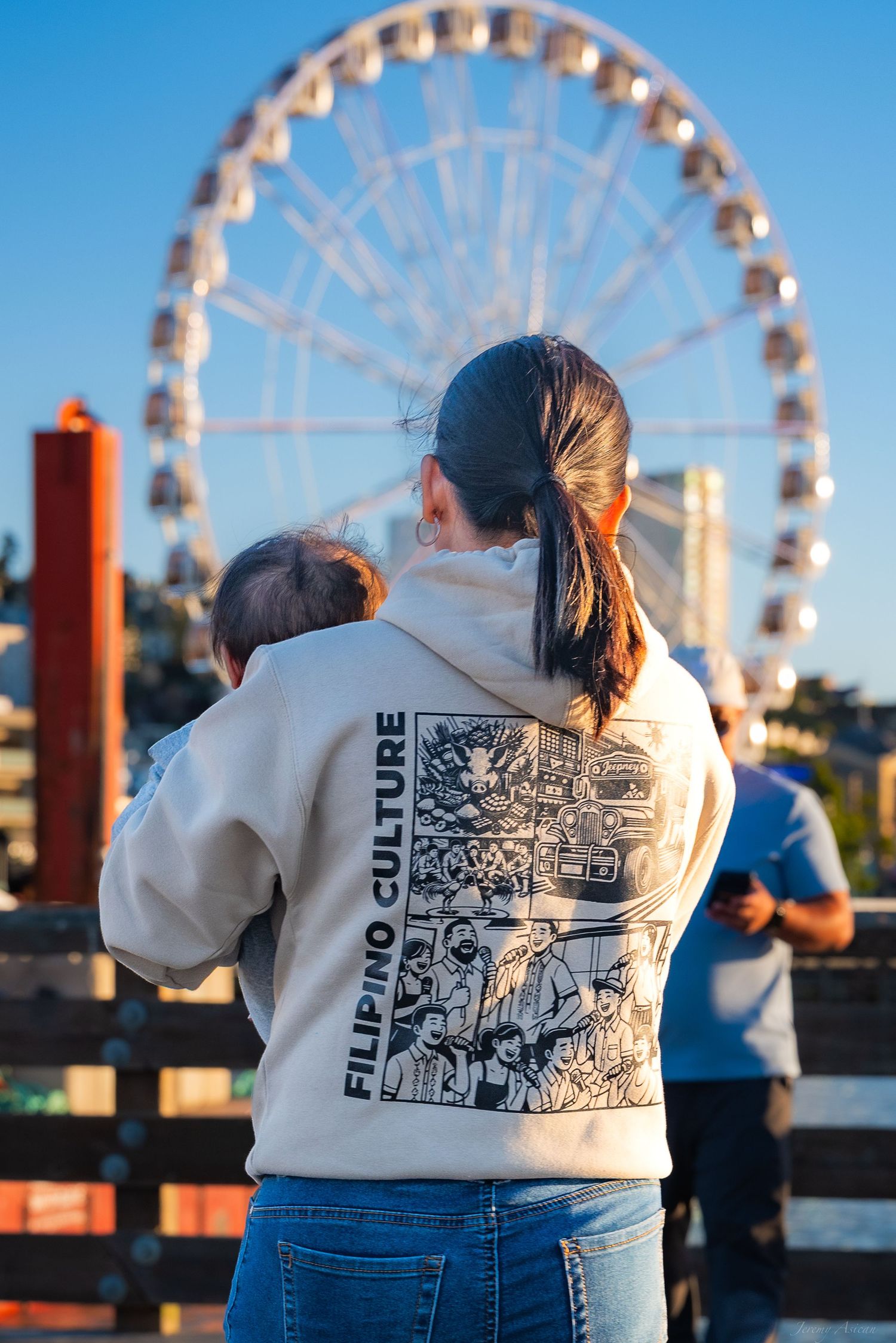 Woman wearing a Filipino Culture hoodie holds a baby while standing in front of a large Ferris wheel at sunset.